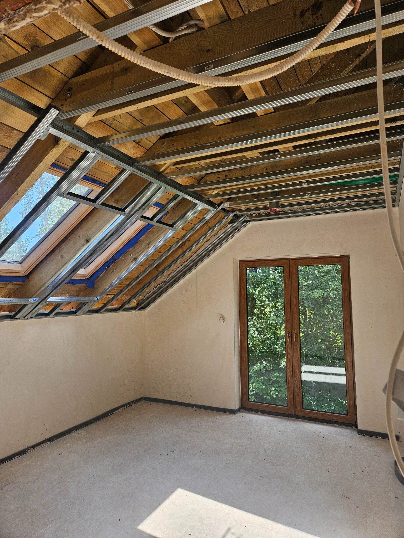 Poddasze w trakcie adaptacji: widoczne drewniane elementy konstrukcyjne dachu, metalowy stelaż pod płyty, okno dachowe i drzwi balkonowe z widokiem na zieleń.