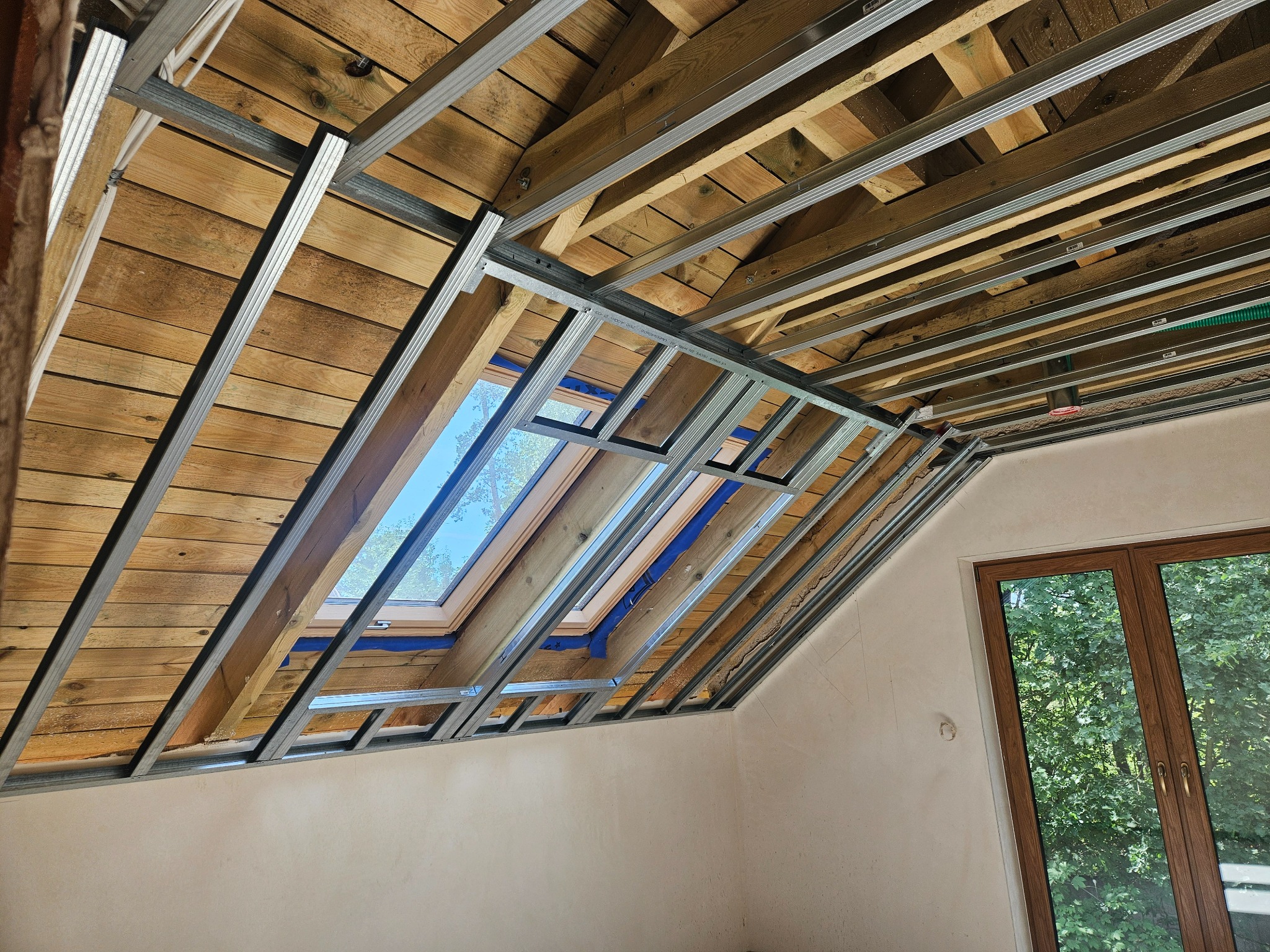 Montaż metalowej konstrukcji pod płytę G-K na drewnianym poddaszu z oknem dachowym. Widoczna konstrukcja i drewniane deski. W tle okno z widokiem na zieleń.