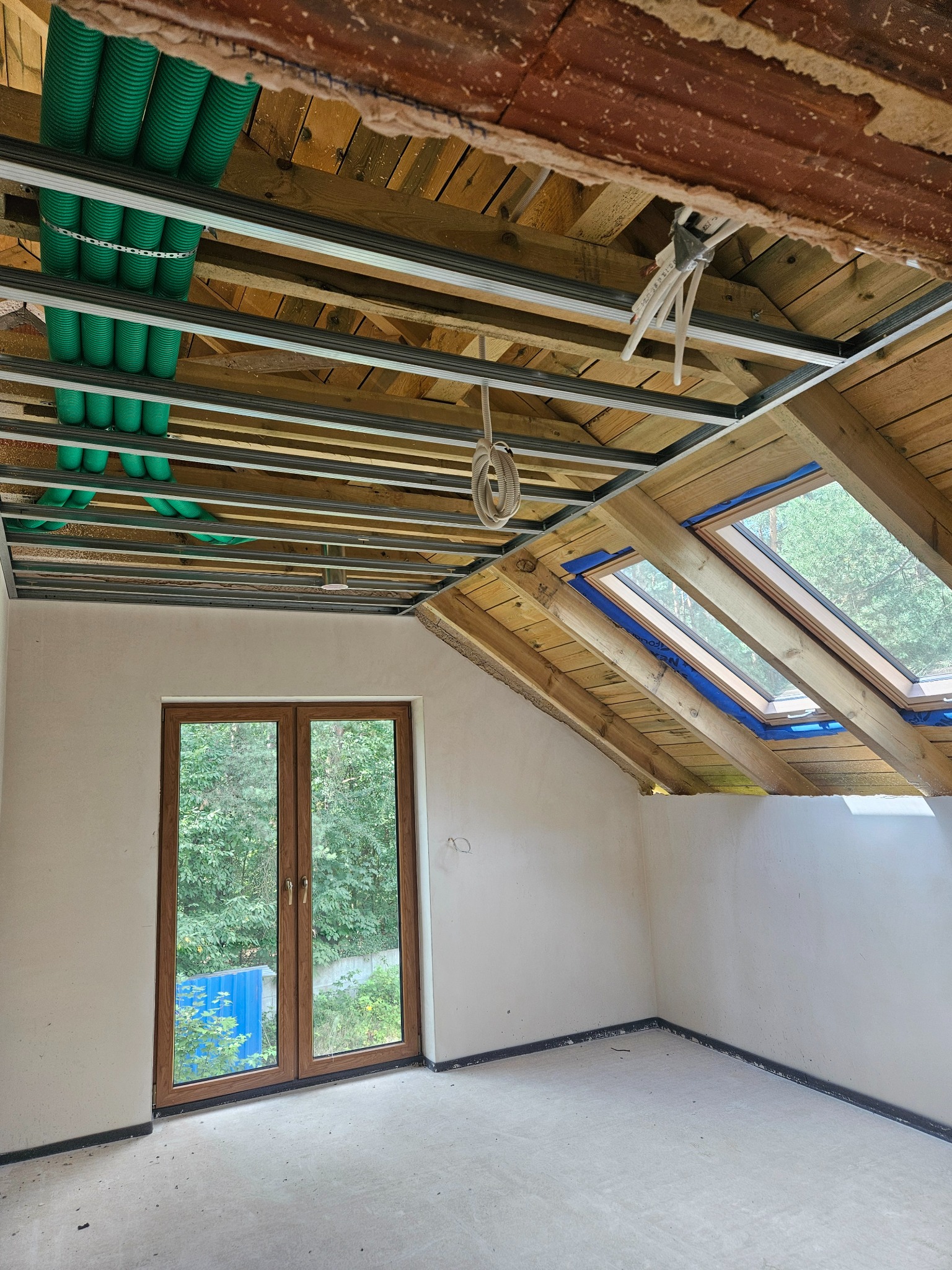 Poddasze w trakcie adaptacji: widoczne okna dachowe, instalacja pod sufitem z rurami wentylacyjnymi i profile do płyt gipsowych. Ściany przygotowane do dalszych prac wykończeniowych.