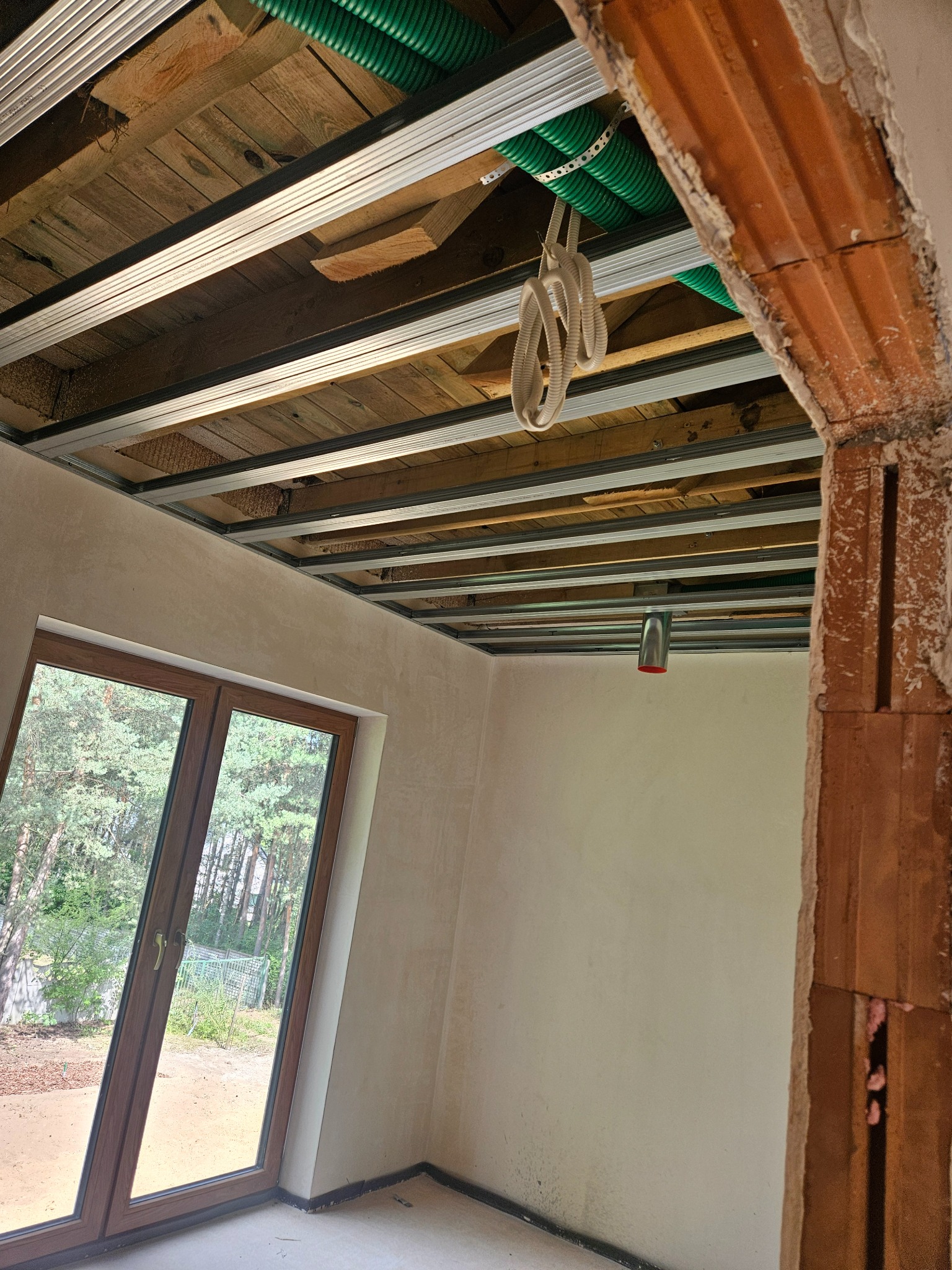 Montaż sufitu podwieszanego z widocznymi instalacjami: rury wentylacyjne i przewody elektryczne, w pomieszczeniu z oknem i surową ścianą. Etap prac wykończeniowych.