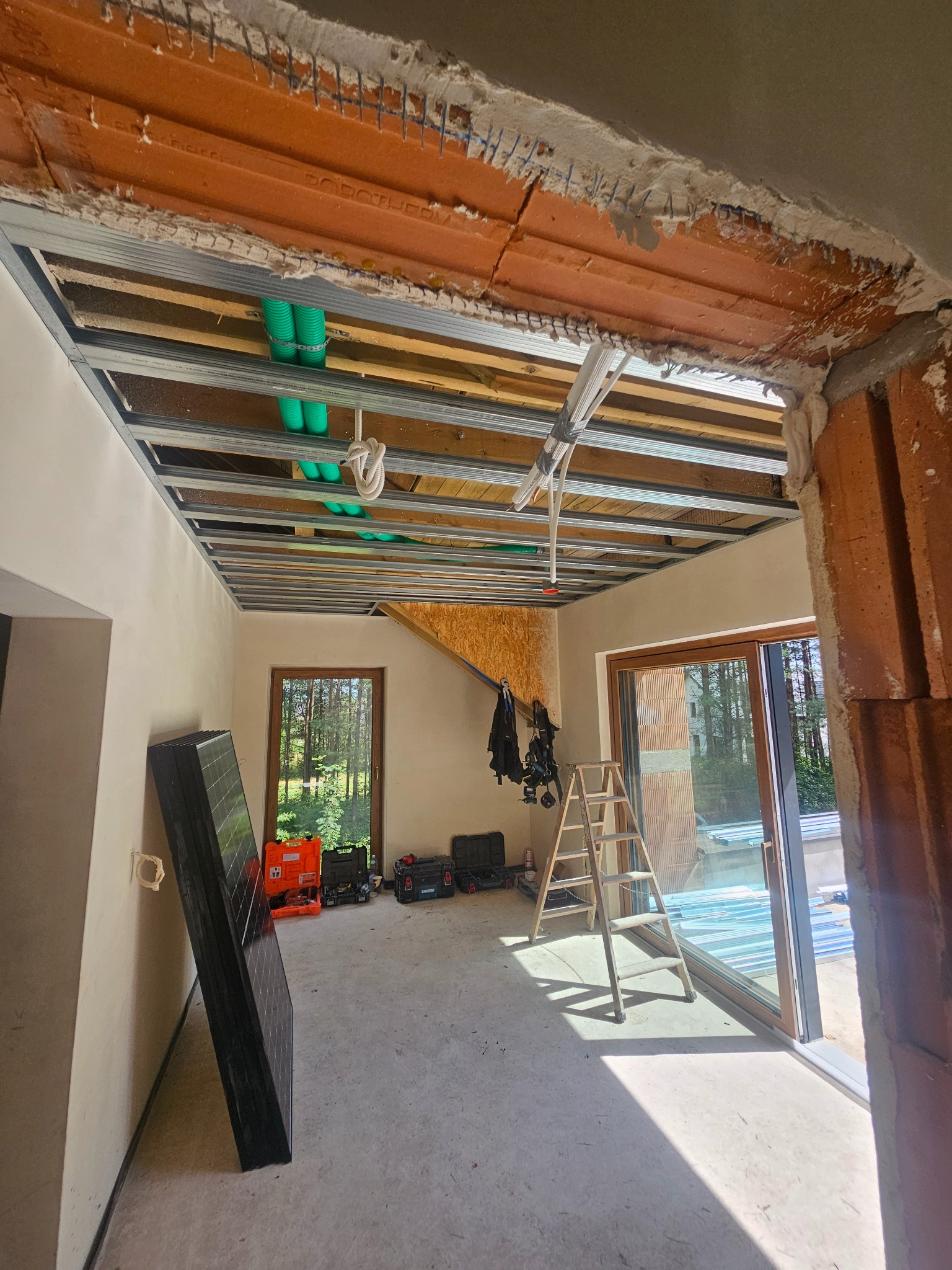 Wnętrze w trakcie montażu sufitu podwieszanego z widocznymi profilami, instalacją i narzędziami. W tle okno z widokiem na zieleń i panele opierające się o ścianę.