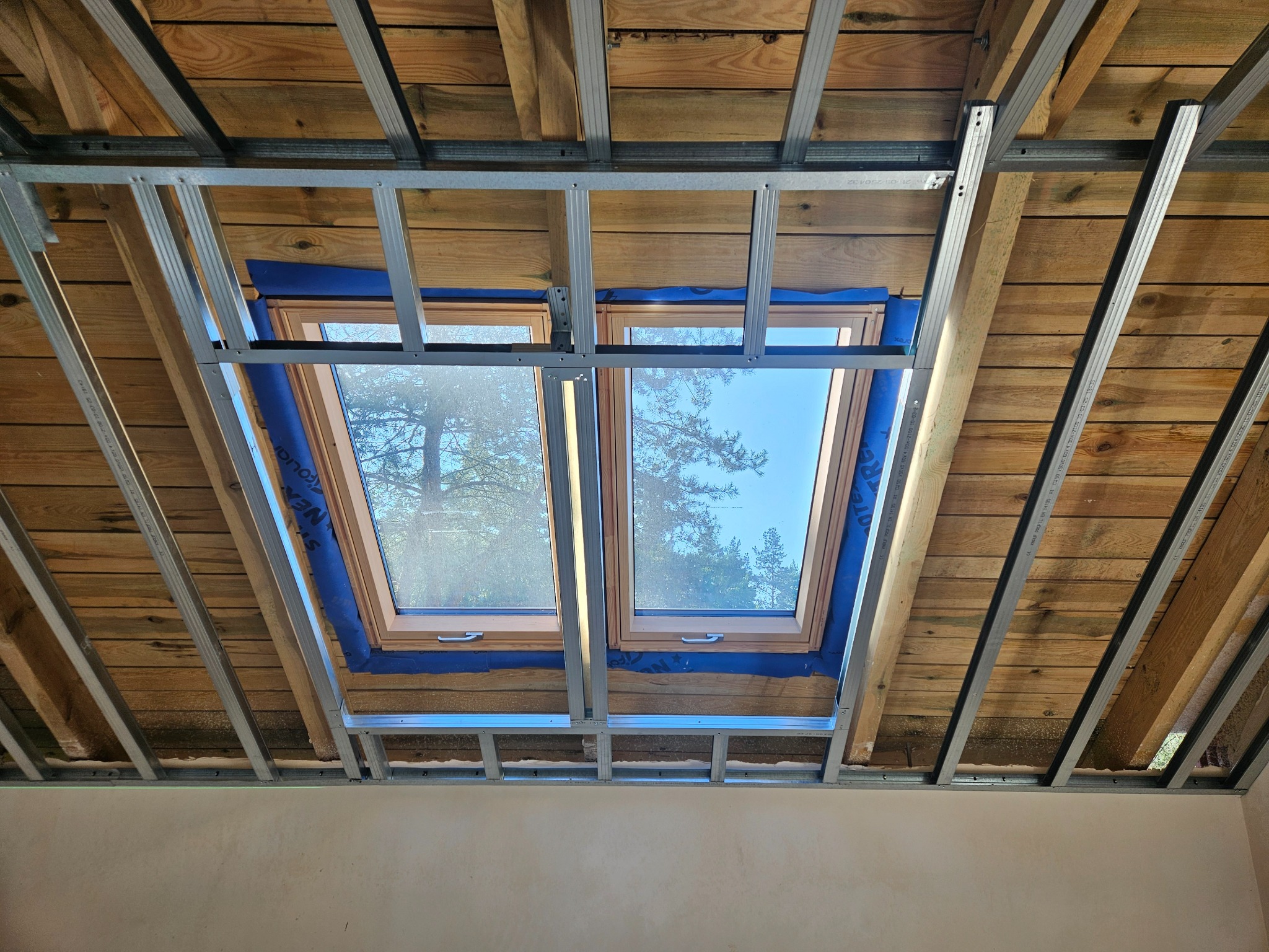 Montaż okien dachowych w drewnianej konstrukcji poddasza z metalowym stelażem, widok z dołu, niebieska folia izolacyjna wokół okien.