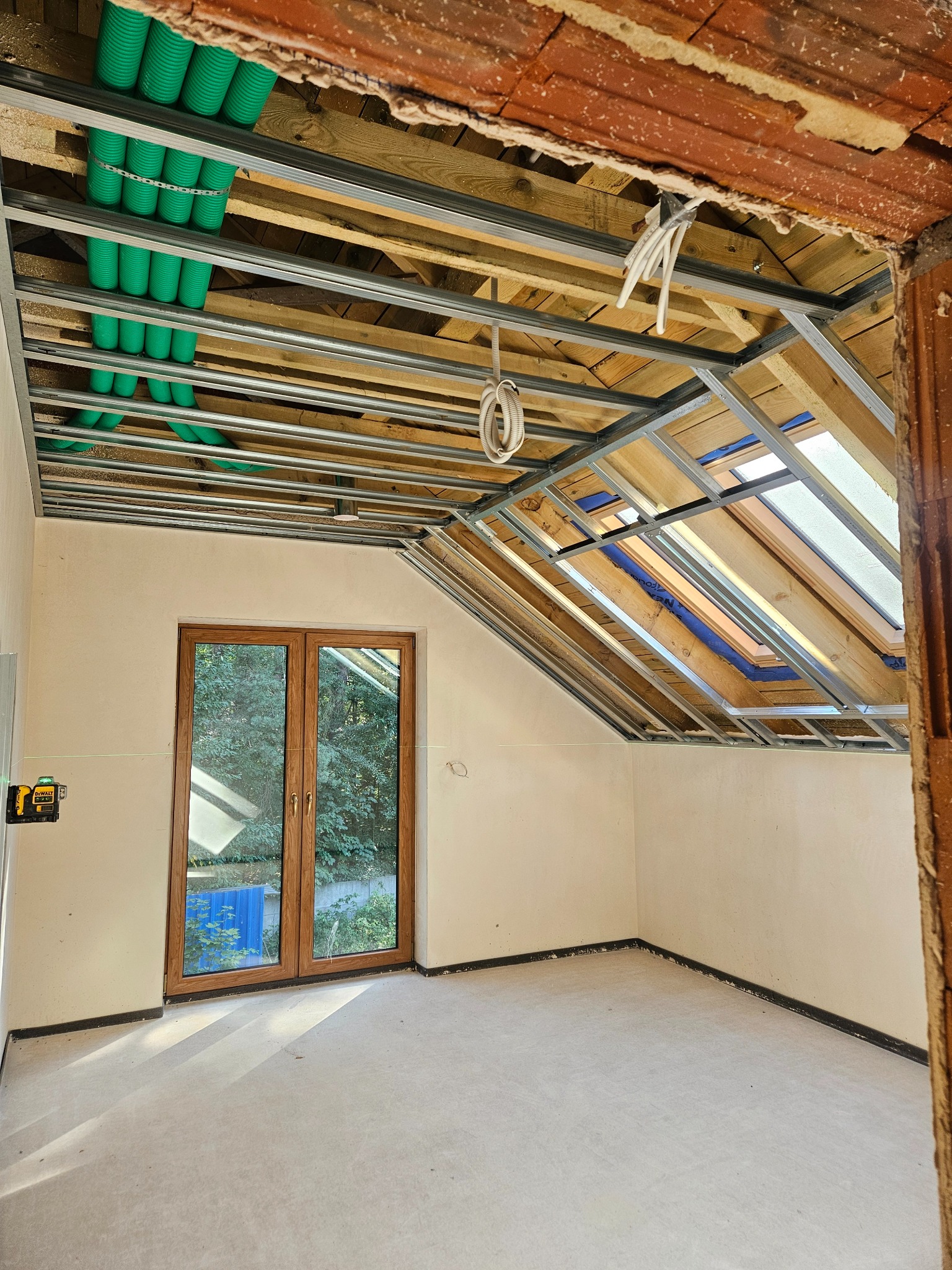 Poddasze w trakcie adaptacji: widoczne metalowe profile sufitowe, zielone rury wentylacyjne, okno dachowe i laser krzyżowy. Przygotowanie do wykończenia.