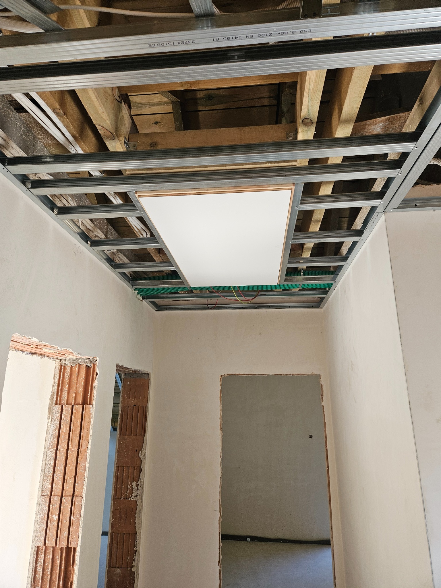 Sufit podwieszany z płytą, widoczne drewniane belki i instalacja elektryczna w zielonej rurze karbowanej. Ściany w trakcie szpachlowania, widoczne otwory drzwiowe.