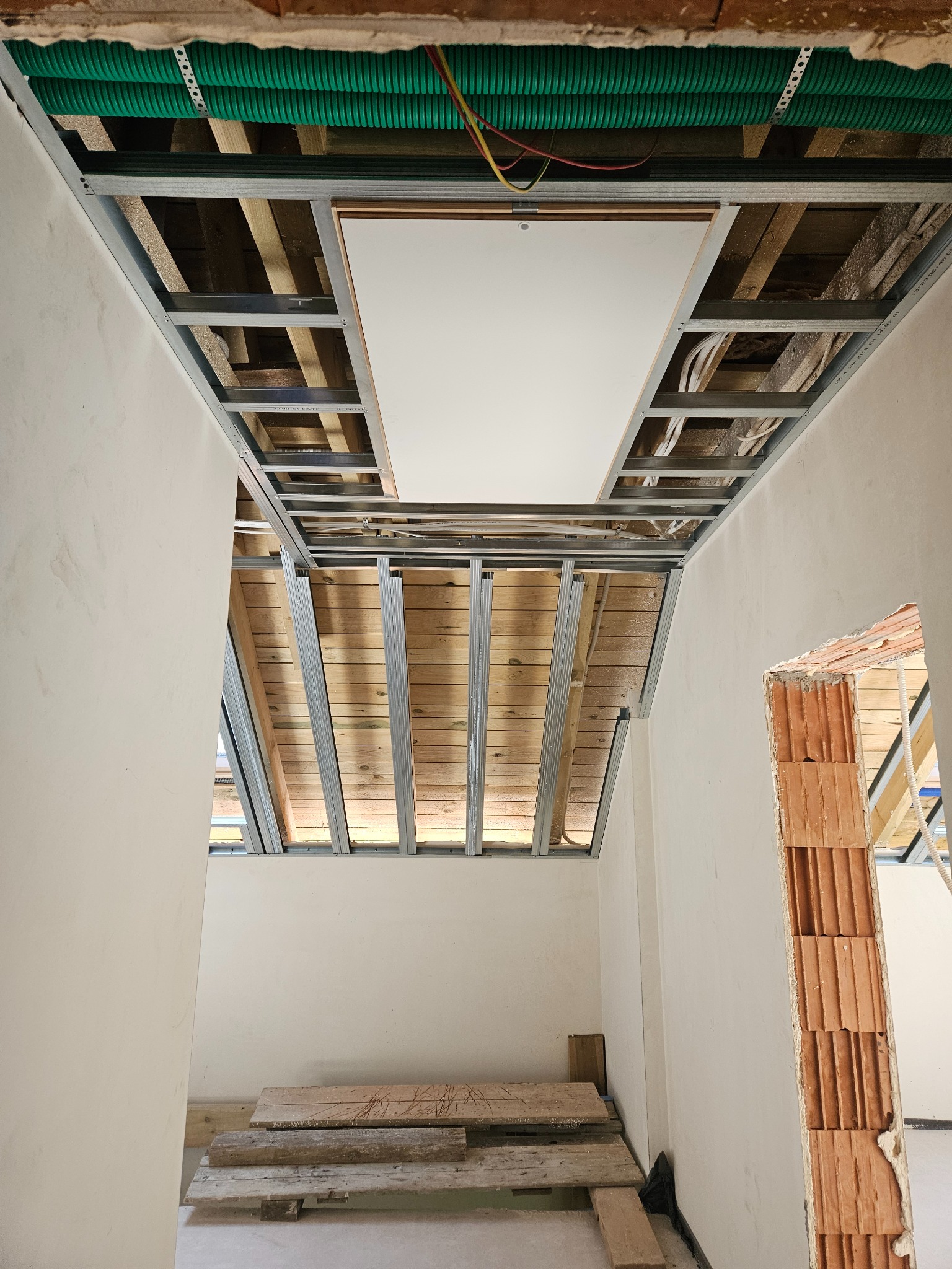 Sufit podwieszany w trakcie montażu, widoczne profile metalowe, instalacje i płyta. W tle ściany w trakcie wykańczania, surowe cegły.