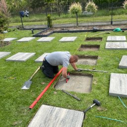 ECO SKILS - Mężczyzna w trakcie układania ścieżki z płyt na trawniku. Widoczne narzędzia, poziomica, szpadel i gumowy młotek. Wykopane otwory pod kolejne płyty.