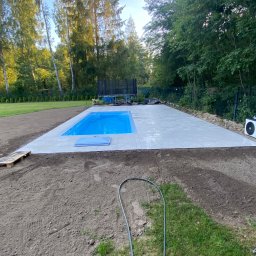 ECO SKILS - Niebieski basen z jasnymi płytkami otoczony ziemią i zielenią. W tle drzewa i trampolina. Widoczne palety i wąż ogrodowy. Kąt ujęcia z dołu.