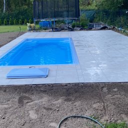 ECO SKILS - Niebieski basen z płytkami w ogrodzie, widok z góry. Trampolina w tle. Świeżo ułożona kostka wokół basenu. Przygotowany teren pod trawnik.