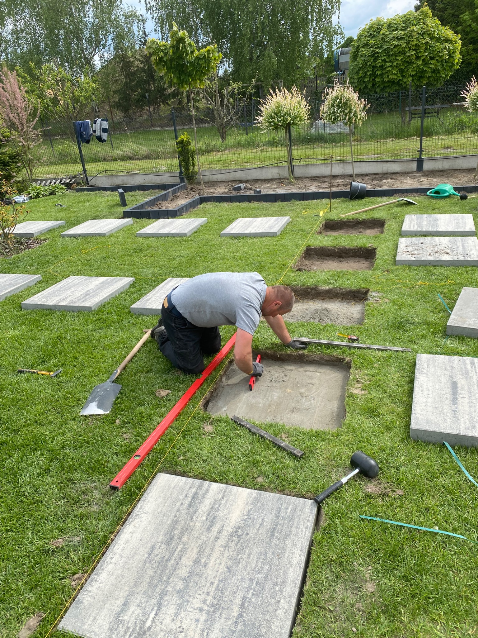 Mężczyzna w trakcie układania ścieżki z płyt na trawniku. Widoczne narzędzia, poziomica, szpadel i gumowy młotek. Wykopane otwory pod kolejne płyty.