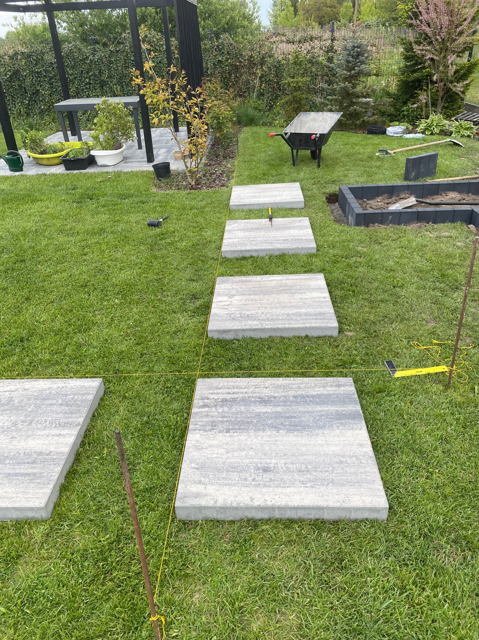 Układanie ścieżki z płyt betonowych na trawniku, widoczne narzędzia i wytyczone linie. Częściowo ukończona realizacja ścieżki ogrodowej z kostki.
