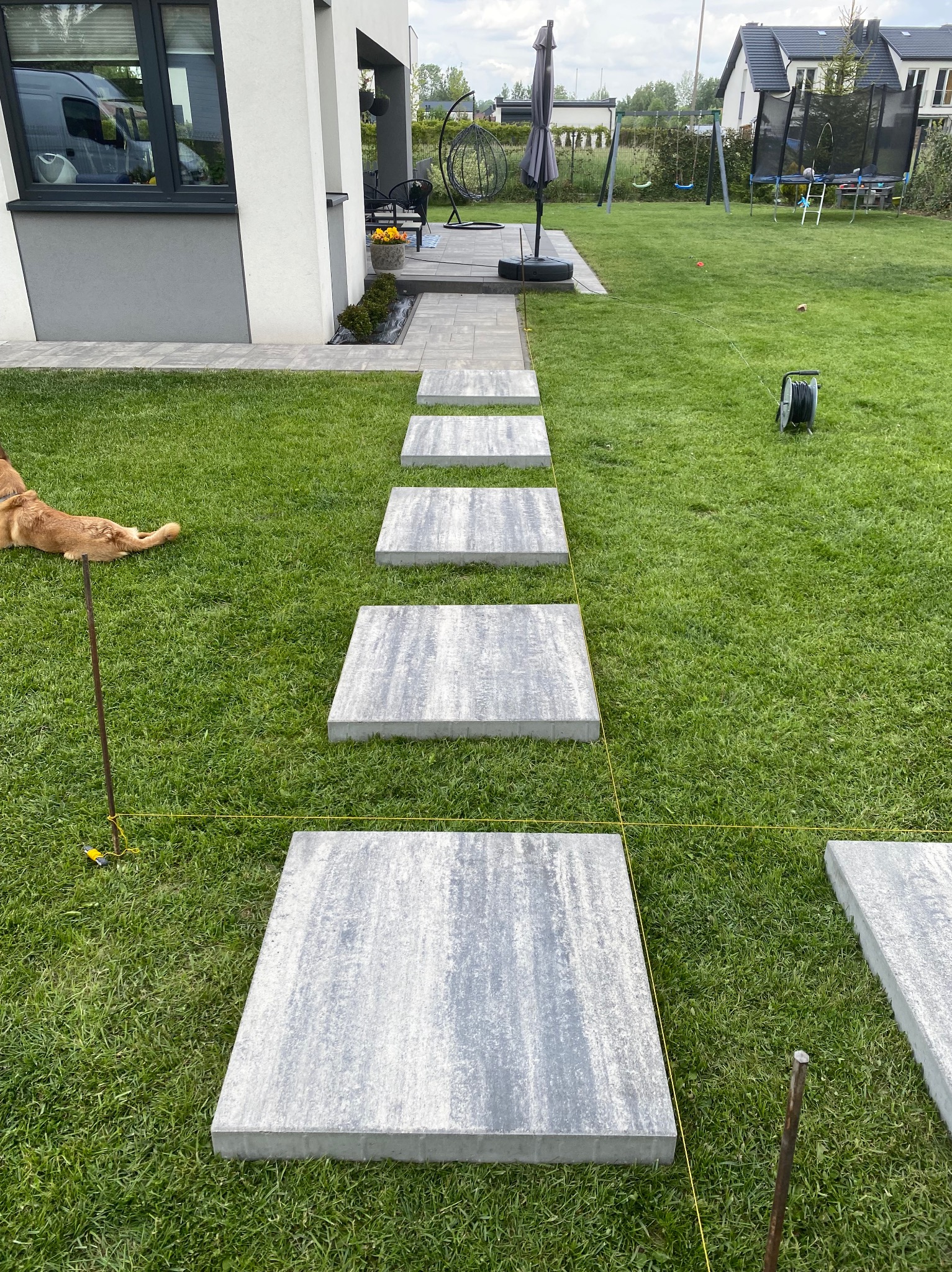Ścieżka z płyt betonowych na trawniku w ogrodzie, wyznaczona żółtą linką, w tle dom z szarą elewacją i huśtawki. Pies leży obok ścieżki.