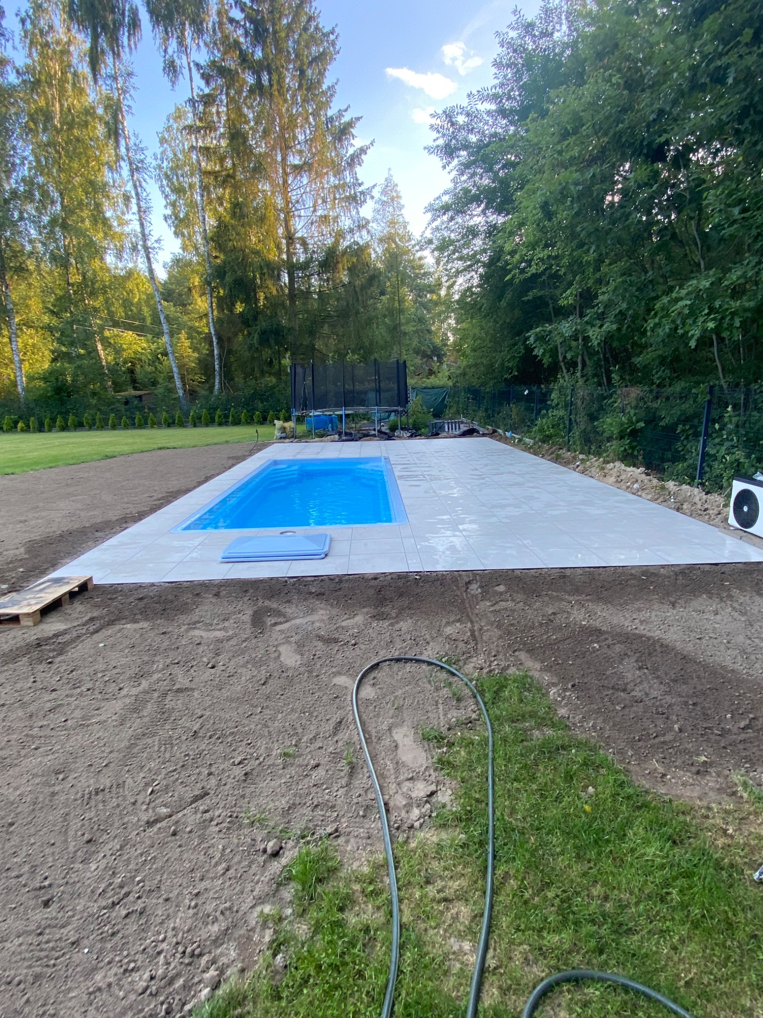 Niebieski basen z jasnymi płytkami otoczony ziemią i zielenią. W tle drzewa i trampolina. Widoczne palety i wąż ogrodowy. Kąt ujęcia z dołu.