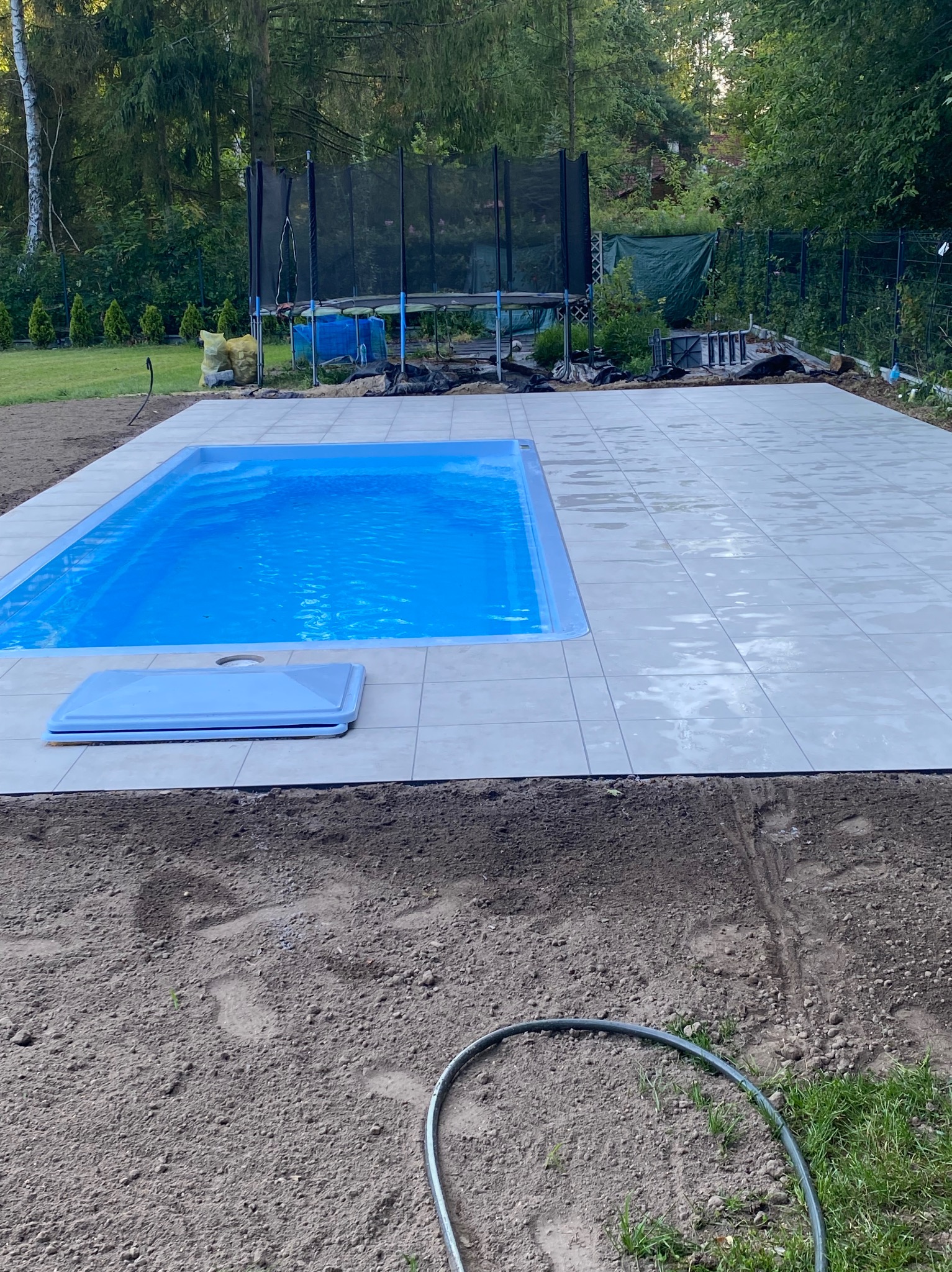 Niebieski basen z płytkami w ogrodzie, widok z góry. Trampolina w tle. Świeżo ułożona kostka wokół basenu. Przygotowany teren pod trawnik.