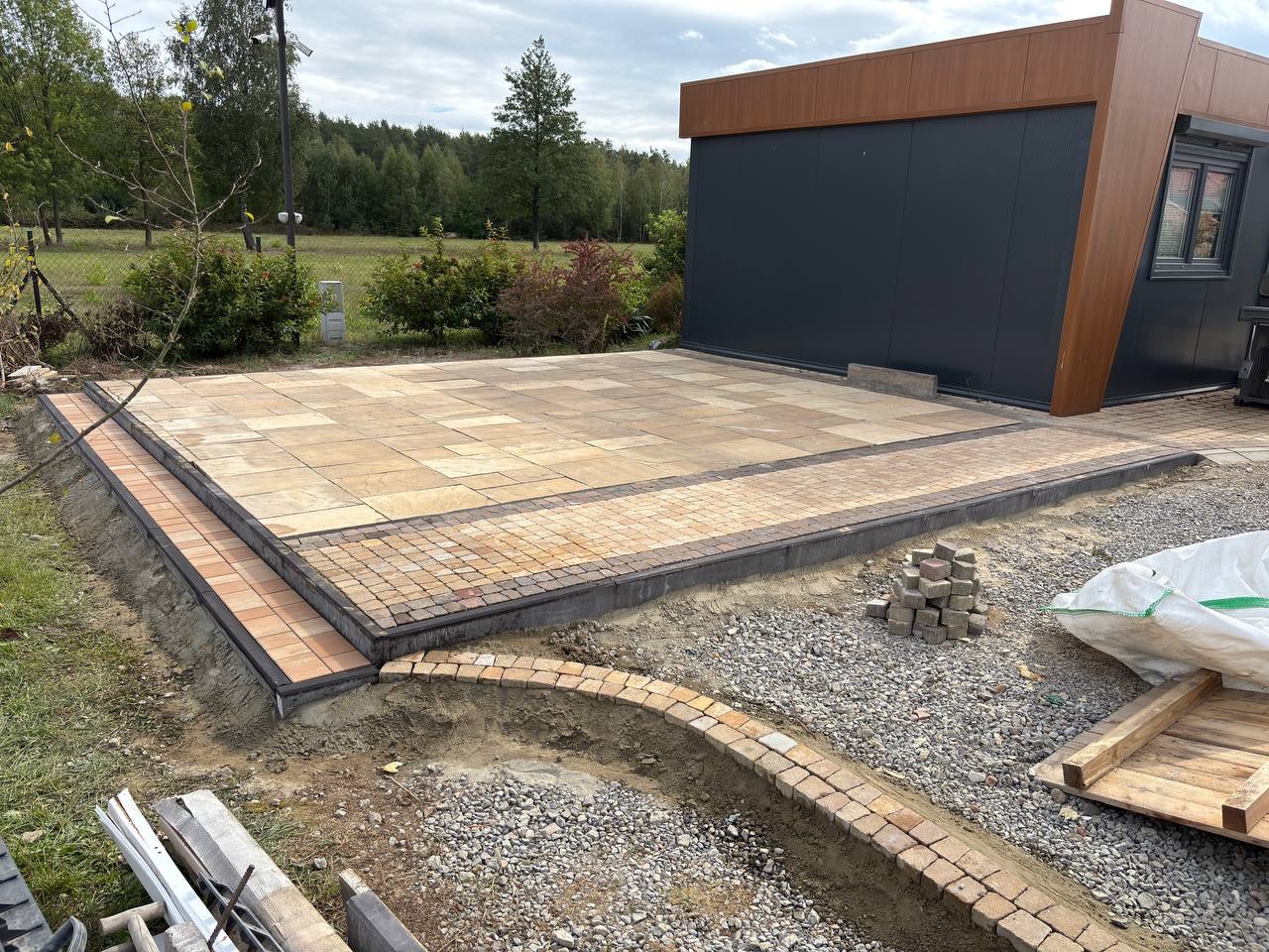 Ułożona kostka brukowa w odcieniach beżu i brązu tworzy plac przy nowoczesnym budynku. Widoczne są fragmenty niezakończonych prac i przygotowane materiały.