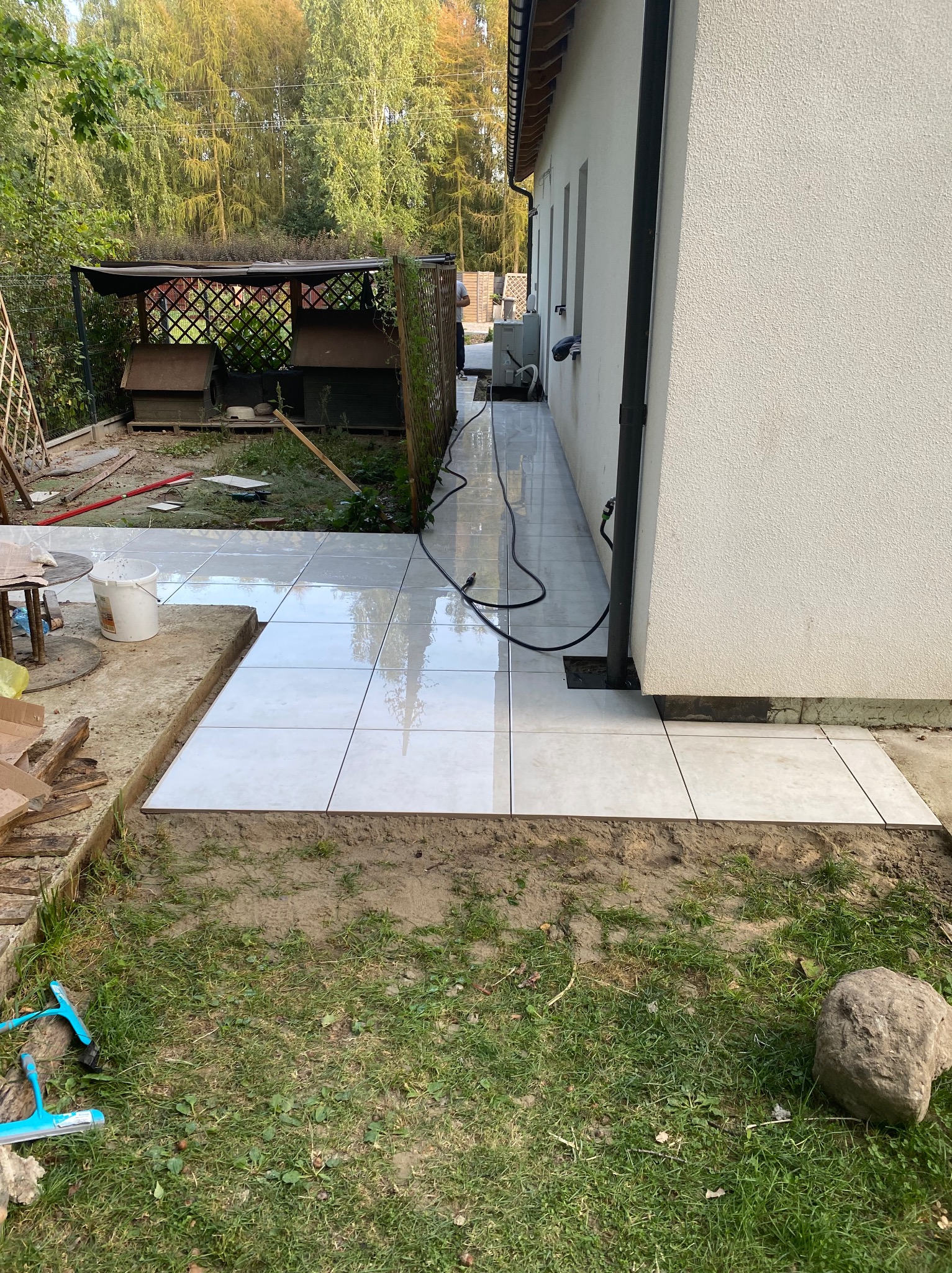 Układanie jasnych płytek chodnikowych wzdłuż białej ściany budynku. Widoczne narzędzia i fragment trawnika. Prace w toku, częściowo ukończona ścieżka.