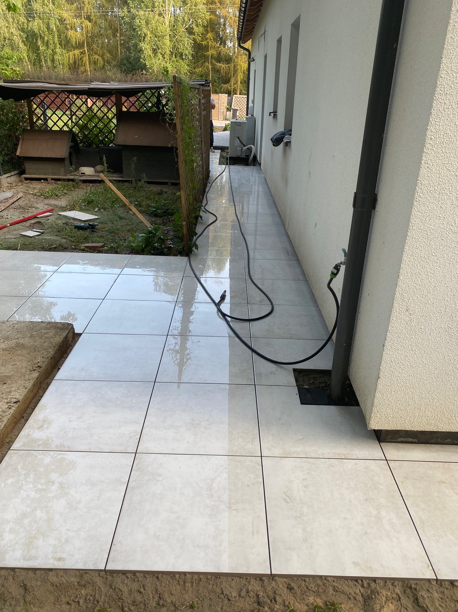 Świeżo ułożone, jasne płytki na zewnątrz budynku, częściowo mokre po myciu. Widoczny wąż ogrodowy i fragment trawnika z budą dla psów w tle.