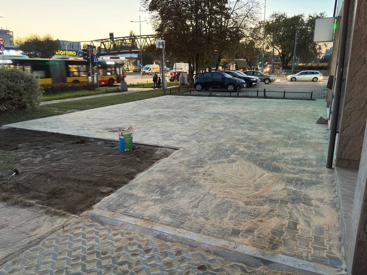 Fragment placu z ułożoną szarą kostką brukową, częściowo zasypaną piaskiem; obok fragment wykopanej ziemi, w tle ulica z samochodami i przejeżdżający autobus.