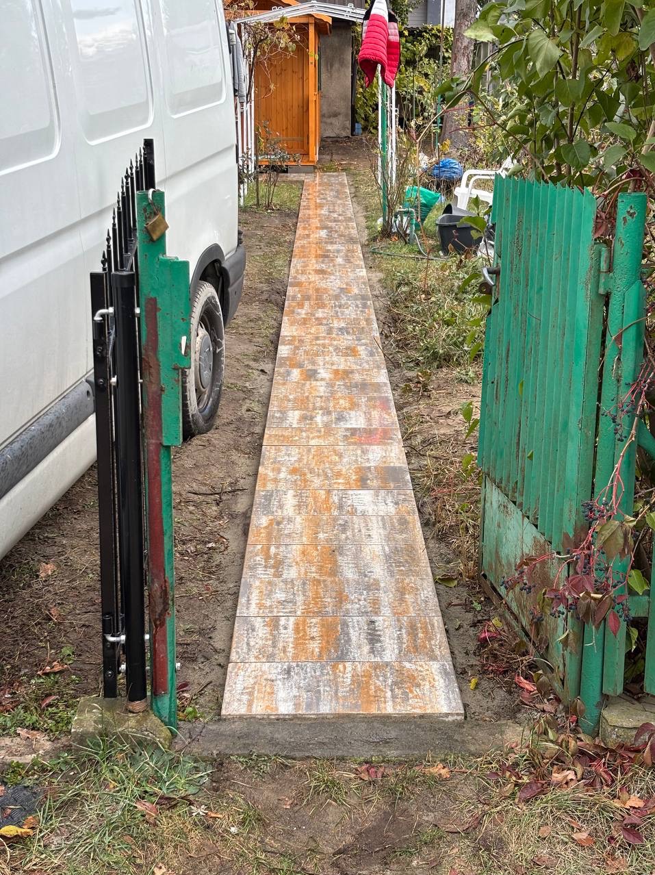 Ułożona ścieżka z kostki brukowej prowadząca od bramki do budynku, widoczna z boku biała furgonetka. Kostka w odcieniach szarości i pomarańczy, otoczenie zarośnięte.