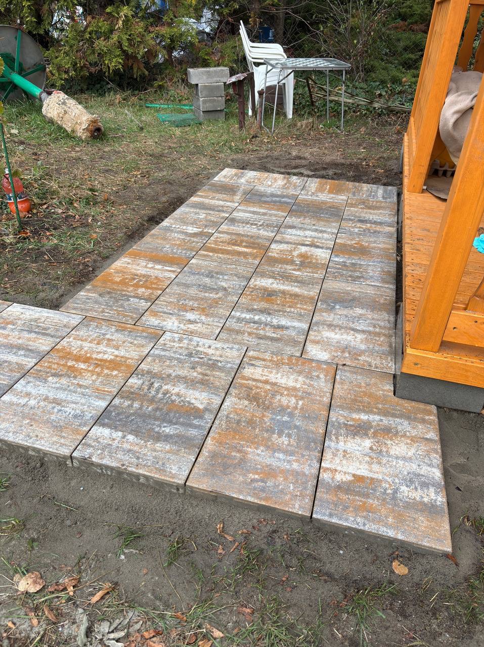 Ułożona ścieżka z kostki brukowej o nieregularnym wzorze, prowadząca do drewnianego podestu. Widoczne elementy ogrodu w tle.