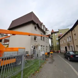 Praca z podnośnika koszowego przy elewacji budynku z brązowym dachem. Widoczne zabezpieczenia, chodnik z kostki i zaparkowany samochód osobowy. Pochmurne niebo.