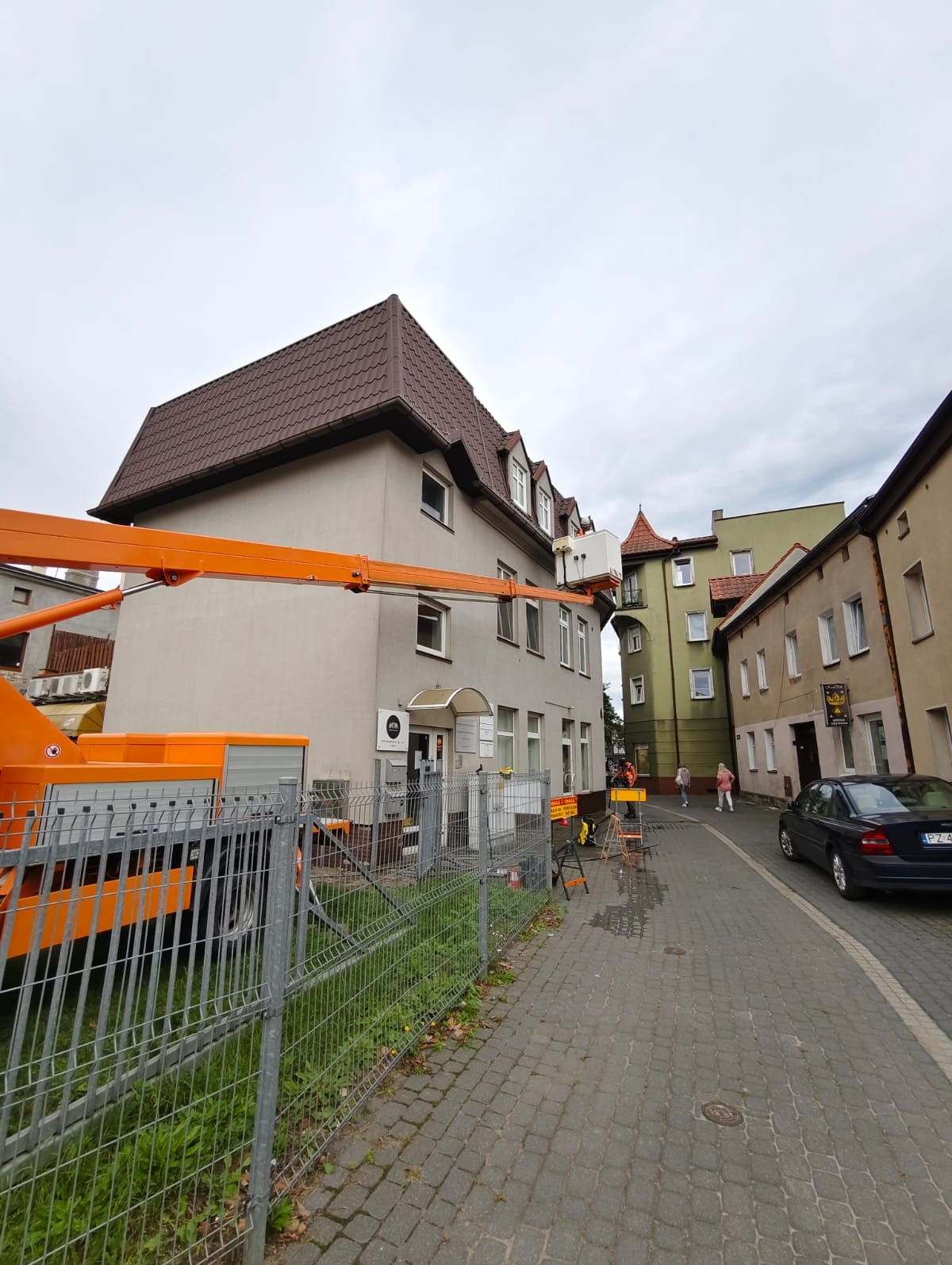 Praca z podnośnika koszowego przy elewacji budynku z brązowym dachem. Widoczne zabezpieczenia, chodnik z kostki i zaparkowany samochód osobowy. Pochmurne niebo.
