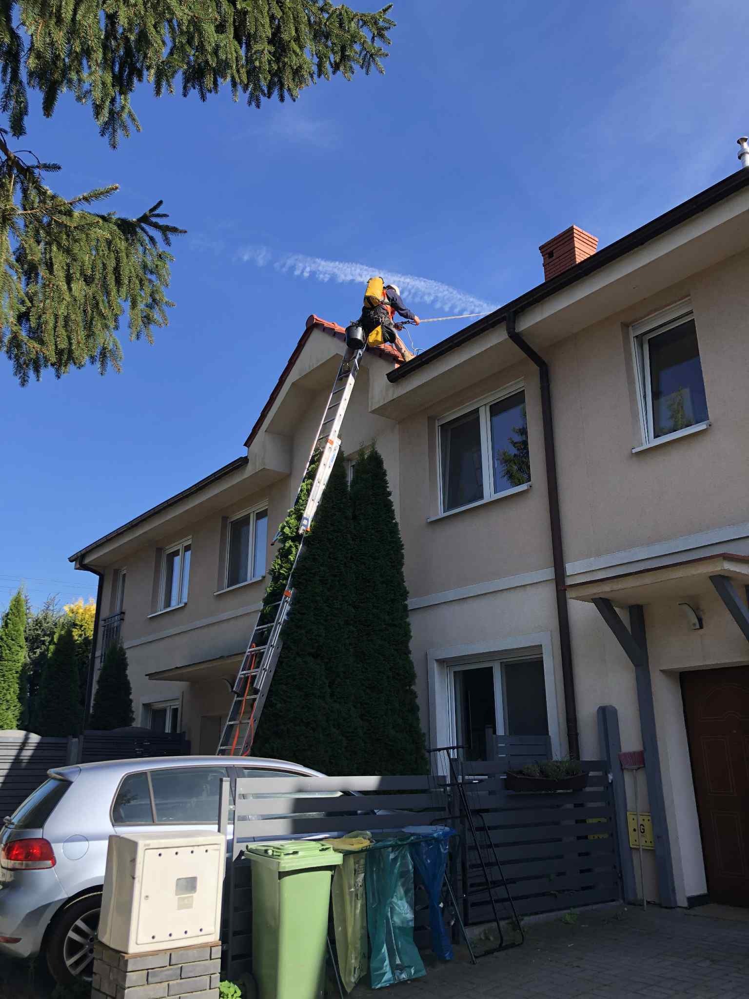 Pracownik na drabinie czyści rynny domu z beżową elewacją w słoneczny dzień. Widoczny samochód i ogrodzenie. Charakterystyczny komin z cegły.