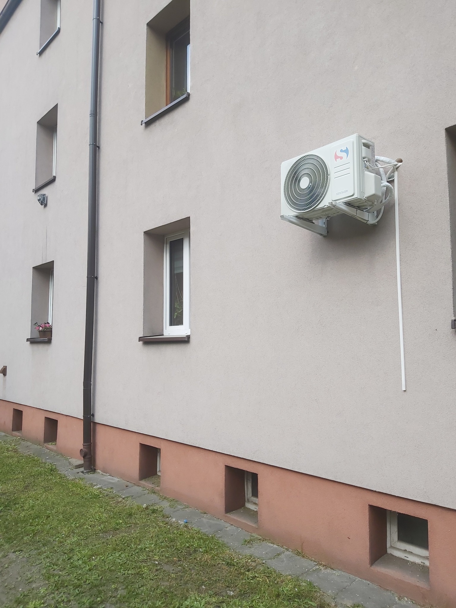 Zamontowany na elewacji budynku klimatyzator Sinclair SKY3 3,2 kW z widocznymi przewodami i elementami instalacji. Ujęcie z dołu, trawnik w tle.