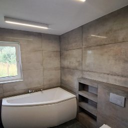 Paweł Kobyliński - Nowoczesna łazienka z narożną wanną, szarymi płytkami i oświetleniem LED. Widok na okno i zabudowę z półkami. Minimalistyczny design.