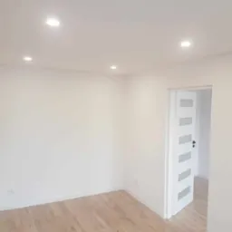 Jasne wnętrze po remoncie: białe ściany, drewnopodobna podłoga, nowoczesne oświetlenie sufitowe i białe drzwi z przeszkleniami.