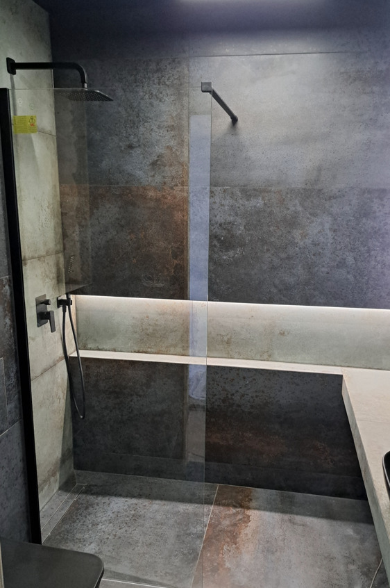 Nowoczesna kabina prysznicowa typu walk-in z czarnymi elementami armatury i szaro-brązowymi płytkami imitującymi beton, podświetlona listwą LED.