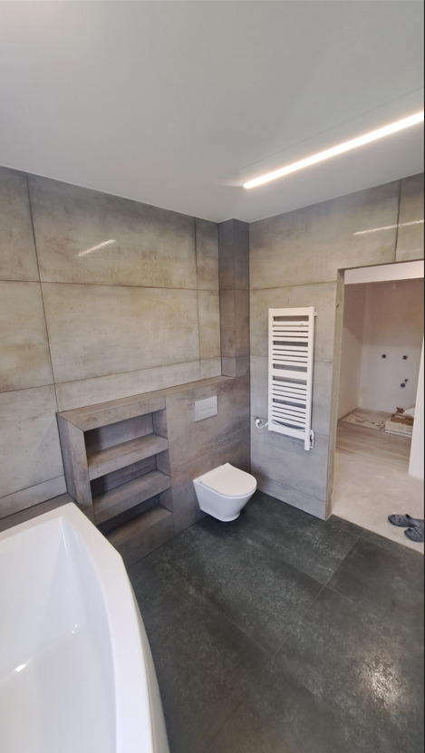 Nowoczesna łazienka z szarymi płytkami wielkoformatowymi, wnęką z półkami i grzejnikiem drabinkowym. W tle widoczny otwarty pokój z podłogą w trakcie prac.