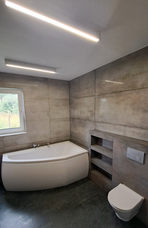 Nowoczesna łazienka z narożną wanną, szarymi płytkami i oświetleniem LED. Widok na okno i zabudowę z półkami. Minimalistyczny design.