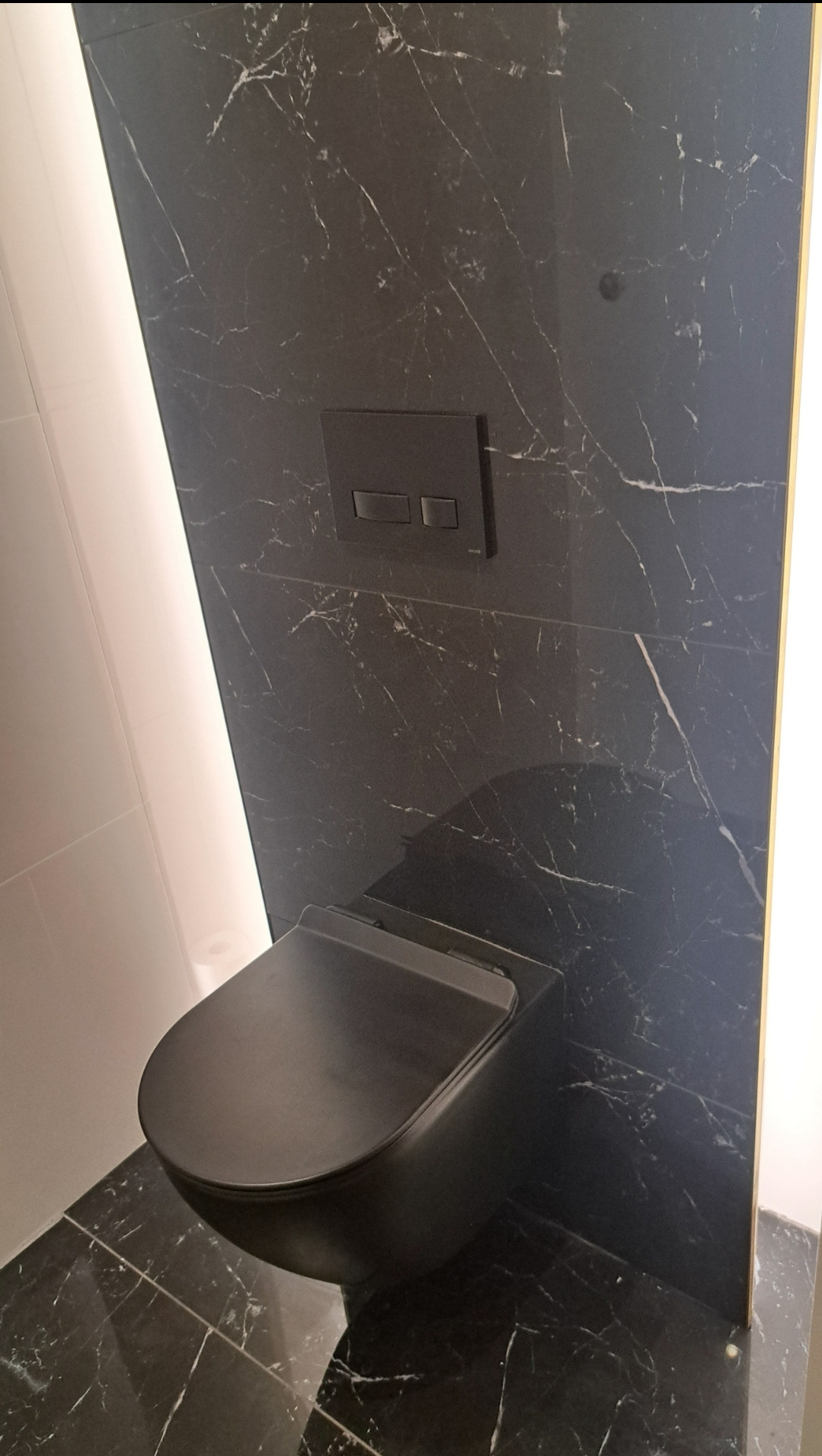 Elegancka, czarna toaleta podwieszana na tle ściany wyłożonej czarnym marmurem z białymi żyłami. Minimalistyczny design, oświetlenie LED po bokach.