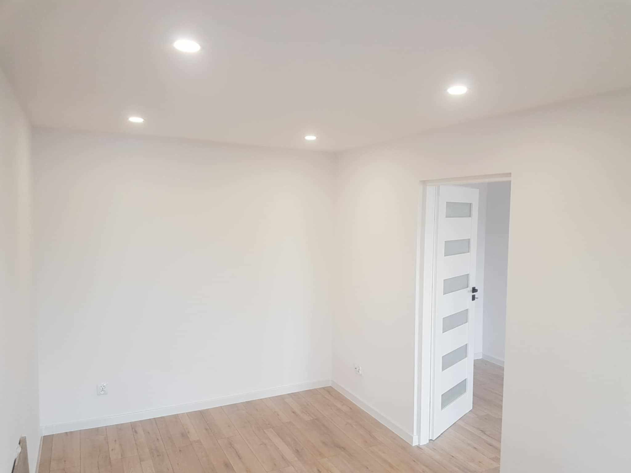 Jasne wnętrze po remoncie: białe ściany, drewnopodobna podłoga, nowoczesne oświetlenie sufitowe i białe drzwi z przeszkleniami.