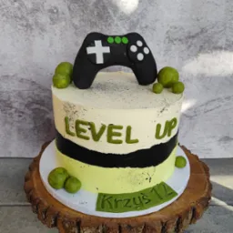 Tort urodzinowy z napisem 'Level Up' i kontrolerem do gier na drewnianym pniu. Dekoracja w odcieniach zieleni, czerni i bieli. Imię 'Krzyś 12' na froncie.