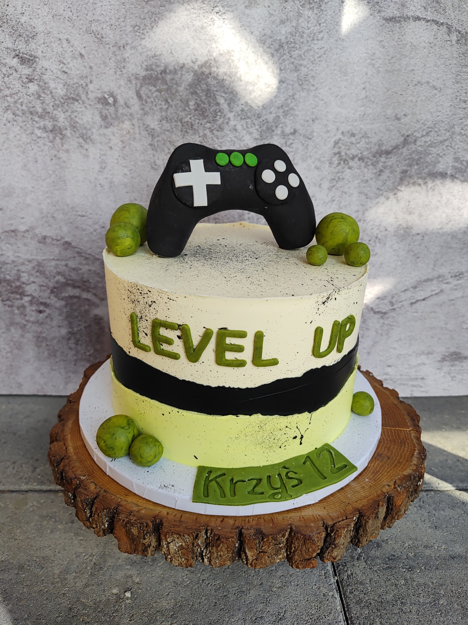 Tort urodzinowy z napisem 'Level Up' i kontrolerem do gier na drewnianym pniu. Dekoracja w odcieniach zieleni, czerni i bieli. Imię 'Krzyś 12' na froncie.