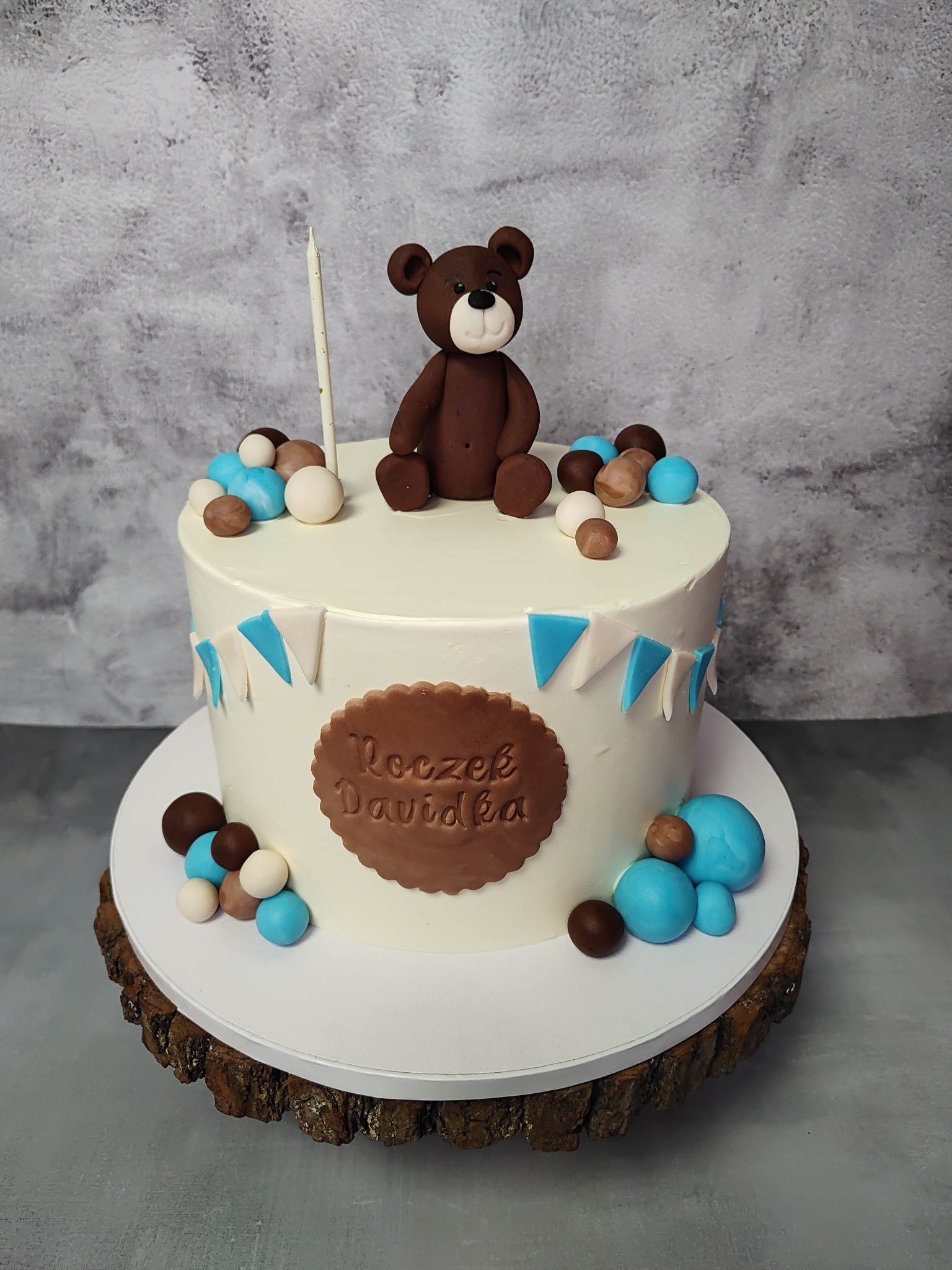 Tort urodzinowy z brązowym misiem na wierzchu, dekorowany niebieskimi i brązowymi kulkami oraz napisem 'Roczek Davidka' na drewnianym podeście.
