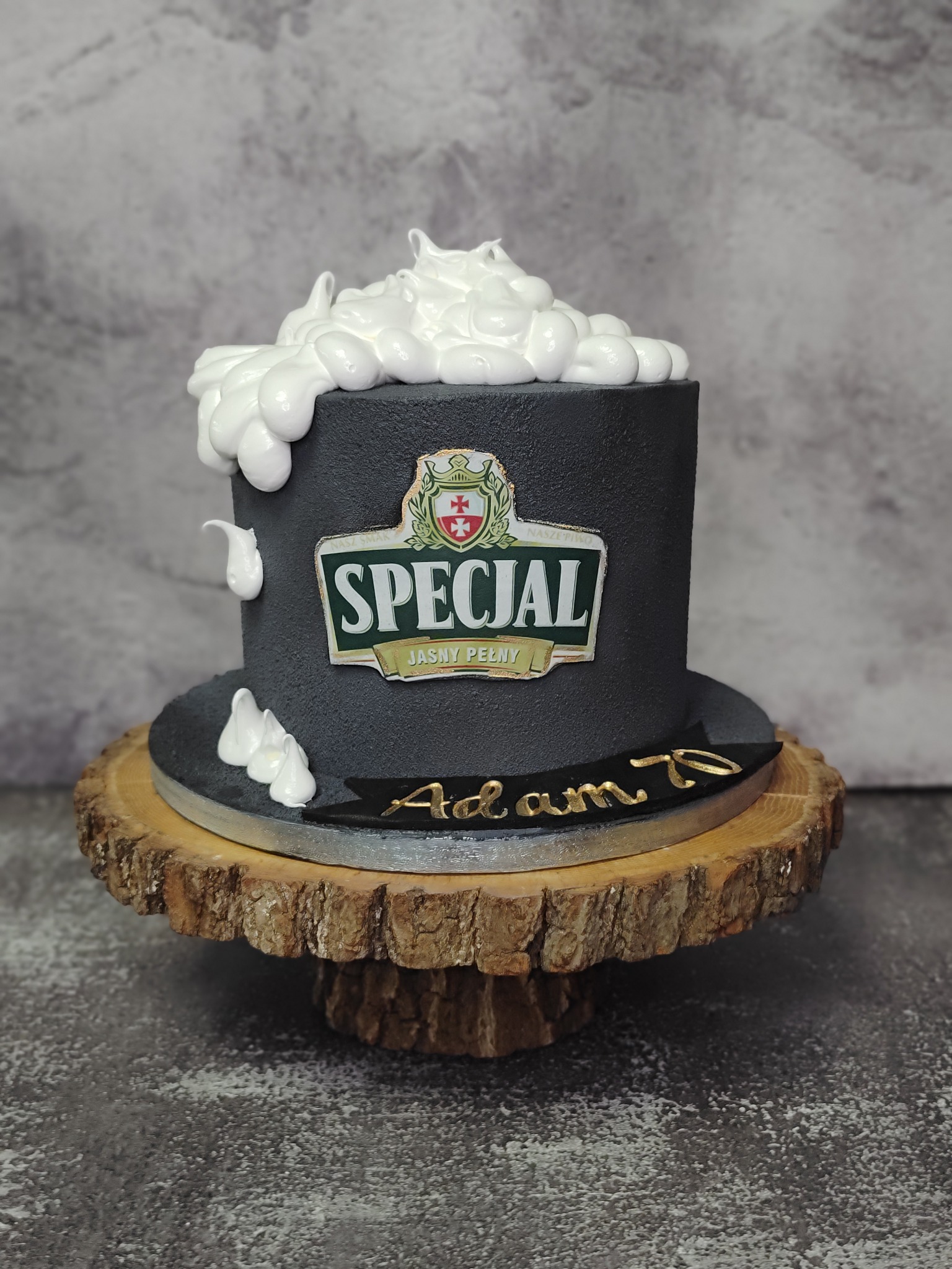 Tort na 70. urodziny z logotypem piwa 'Specjal' i białą pianą imitującą piwo, umieszczony na drewnianym plastrze. Szaro-betonowe tło.