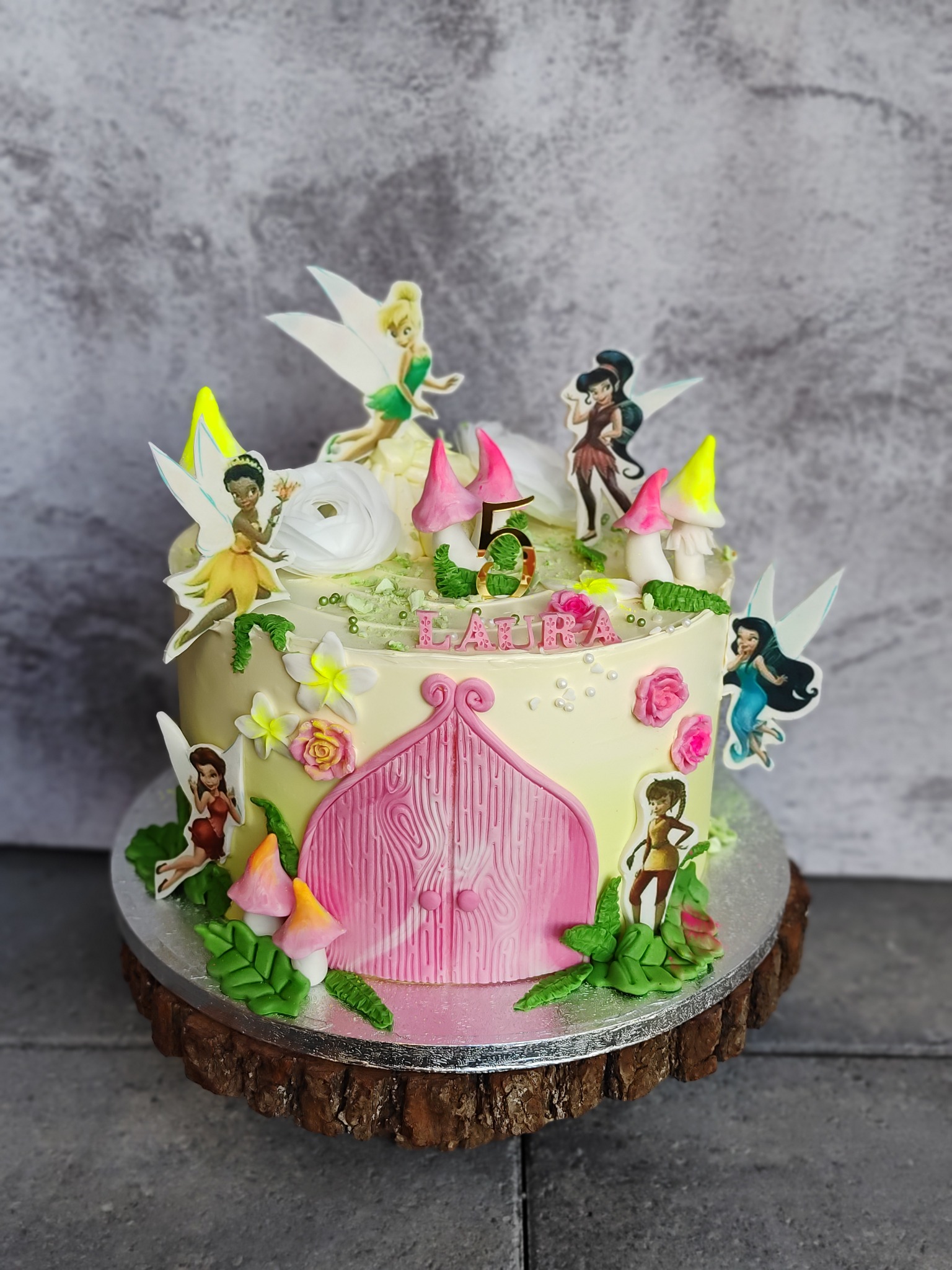 Udekorowany tort urodzinowy z motywem wróżek, różową bramą i napisem 'Laura' na drewnianym stojaku, na tle szarej ściany. Detal cukierniczy, jasne kolory.