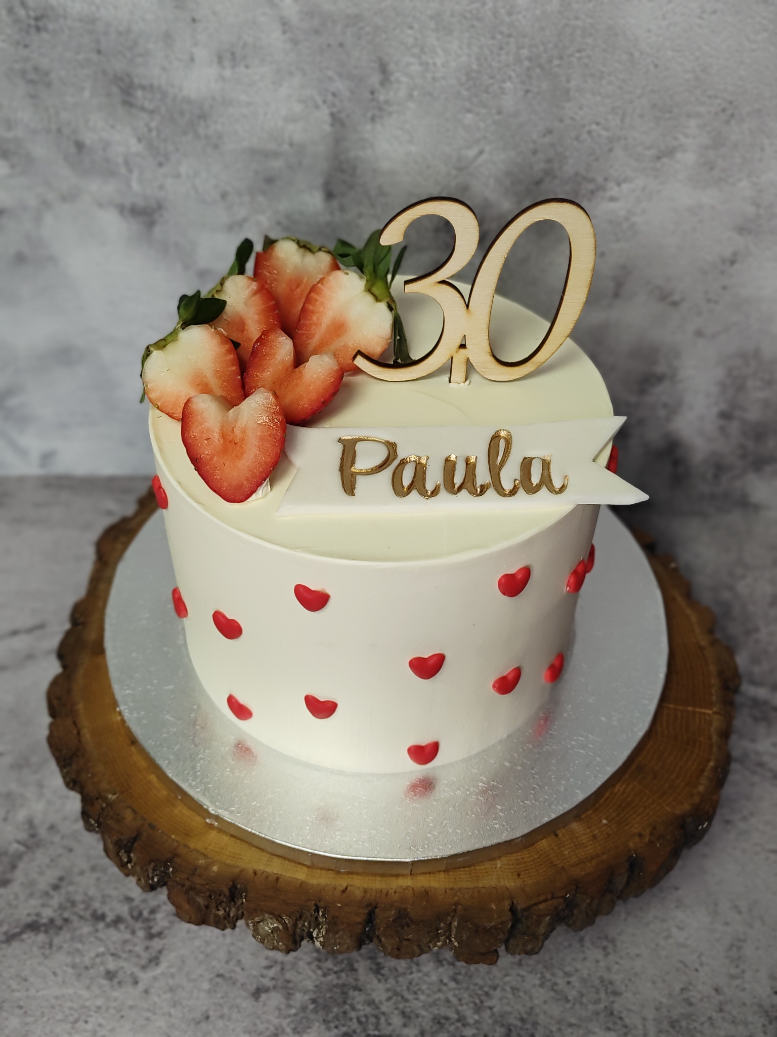 Tort urodzinowy dla Pauli na 30 urodziny. Biały krem, czerwone serduszka, truskawki i drewniany napis. Stoi na drewnianym plastrze.
