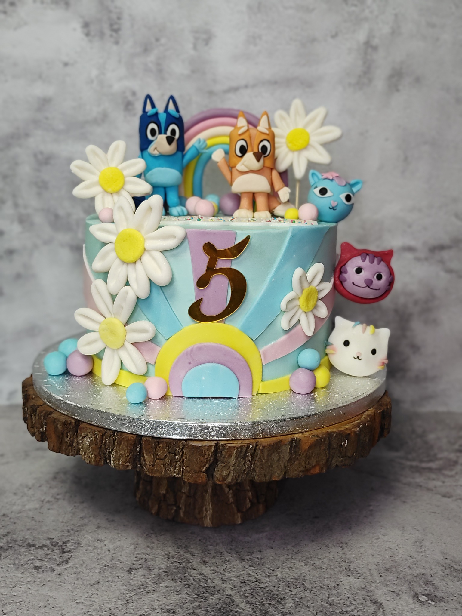 Tort urodzinowy z motywem bajkowym: figurki piesków Bluey i Bingo, tęcza, kwiaty, cyfra 5. Pastelowe kolory, drewniany stojak. Idealny na dziecięce przyjęcie.
