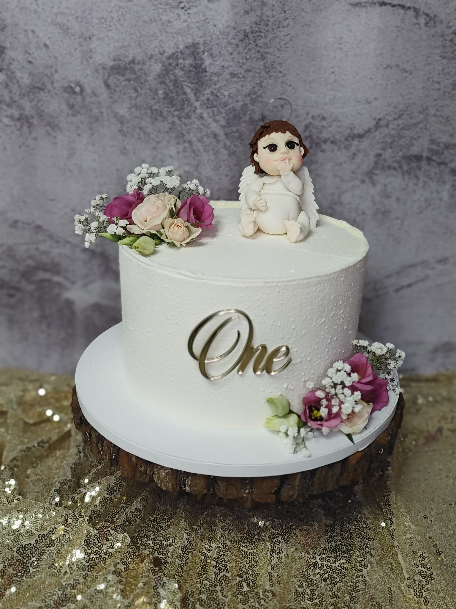 Tort 'One' z figurką aniołka i kwiatami na drewnianym postumencie na tle błyszczącej tkaniny. Idealny na roczek dziecka, wykonany przez cukiernię.