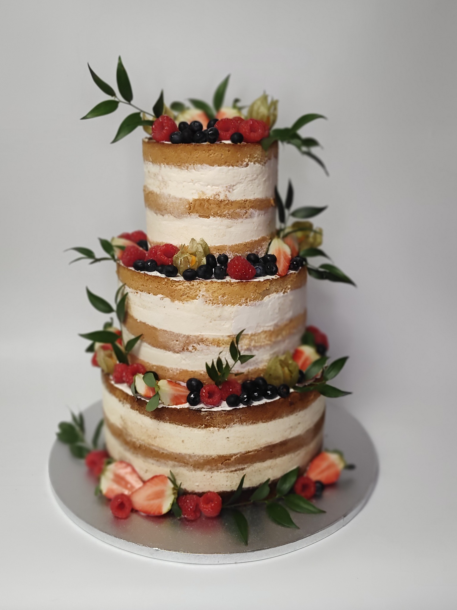 Trzypiętrowy tort 'naked cake' na srebrnej paterze, udekorowany świeżymi owocami (maliny, jagody, truskawki) i zielonymi listkami. Kremowy, delikatny wygląd.