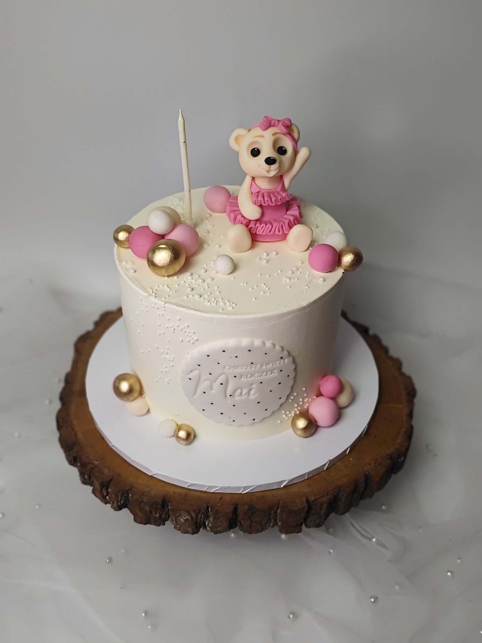 Tort urodzinowy z misiem w różowej sukience, białym kremem i złotymi kulkami, na drewnianym podkładzie. Świeczka na górze. Napis 'Już świętuje roczek'.