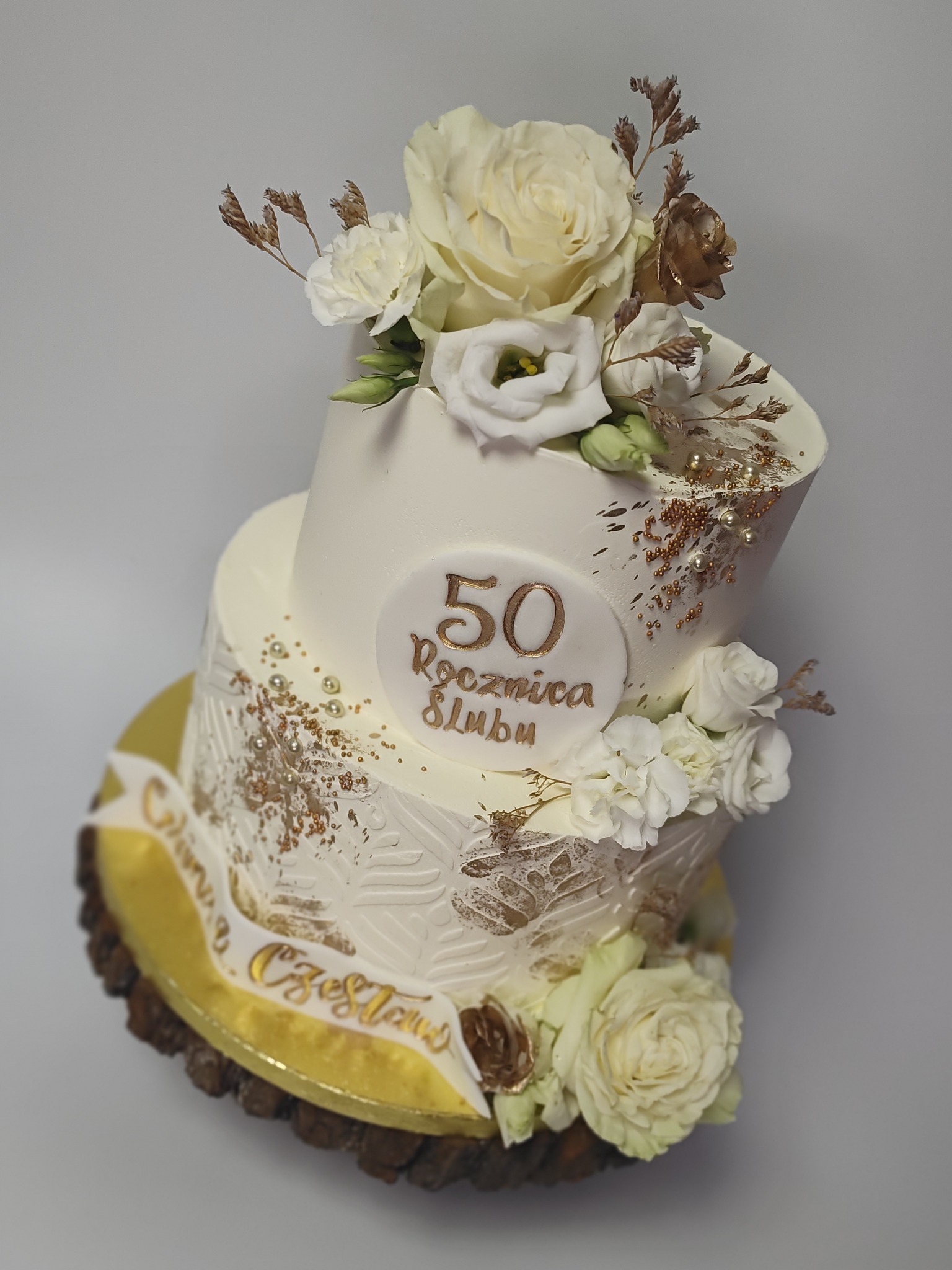 Dwupoziomowy tort na 50. rocznicę ślubu, zdobiony białymi kwiatami, złotymi akcentami i napisem okolicznościowym. Ustawiony na drewnianej podstawie.