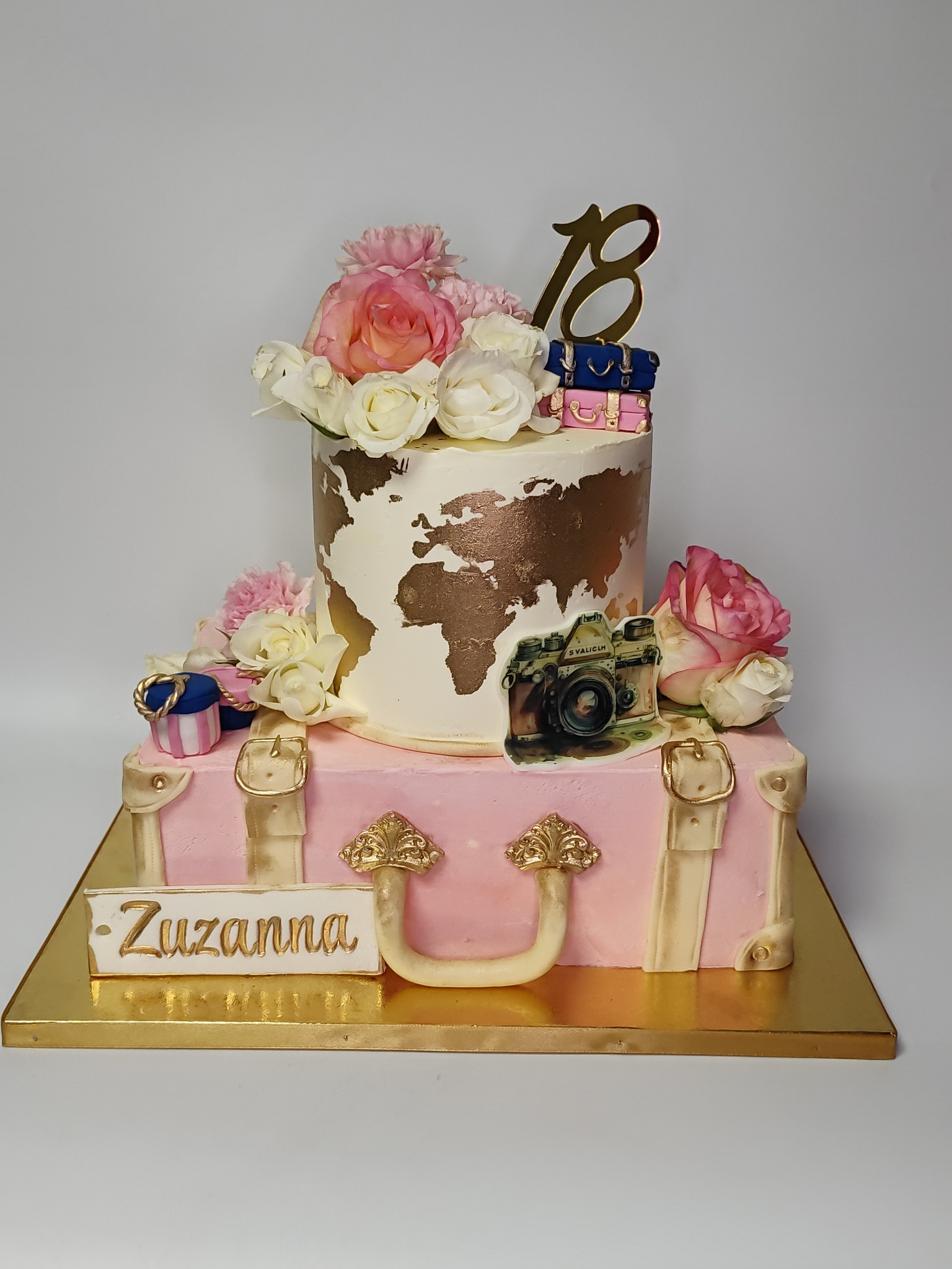 Dwupoziomowy tort w stylu podróżniczym: walizka z imieniem Zuzanna, globus, kwiaty, aparat i cyfra 18. Dekoracja urodzinowa z dbałością o detale.