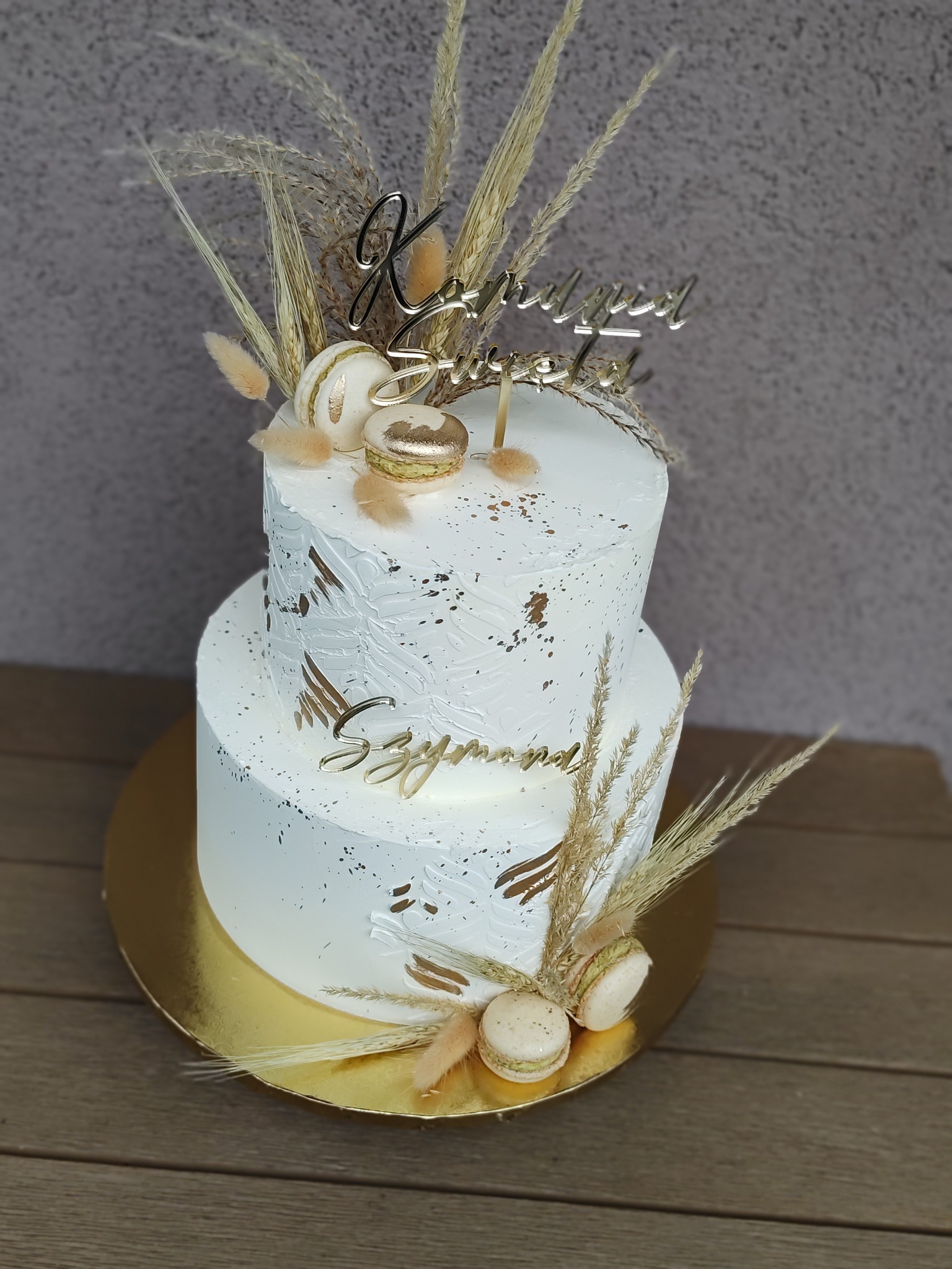 Dwupoziomowy tort w białym kremie, zdobiony złotymi akcentami, makaronikami i suszonymi trawami. Na górze napis 'Kornelia i Szymon'. Elegancki i nowoczesny.
