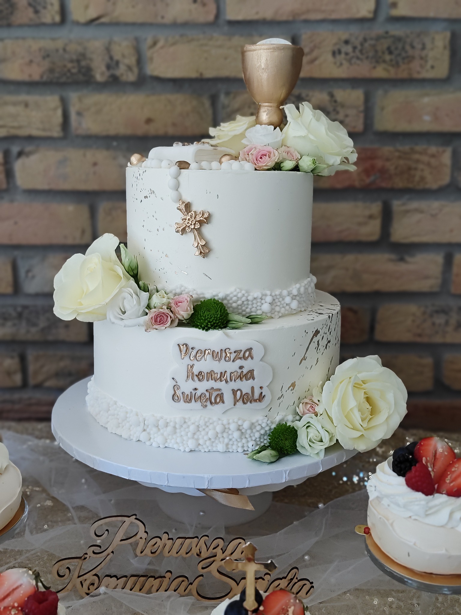 Dwupiętrowy tort komunijny w bieli, zdobiony złotym kielichem, krzyżem i białymi różami, na tle ceglanej ściany. Napis 'Pierwsza Komunia Święta Poli'.