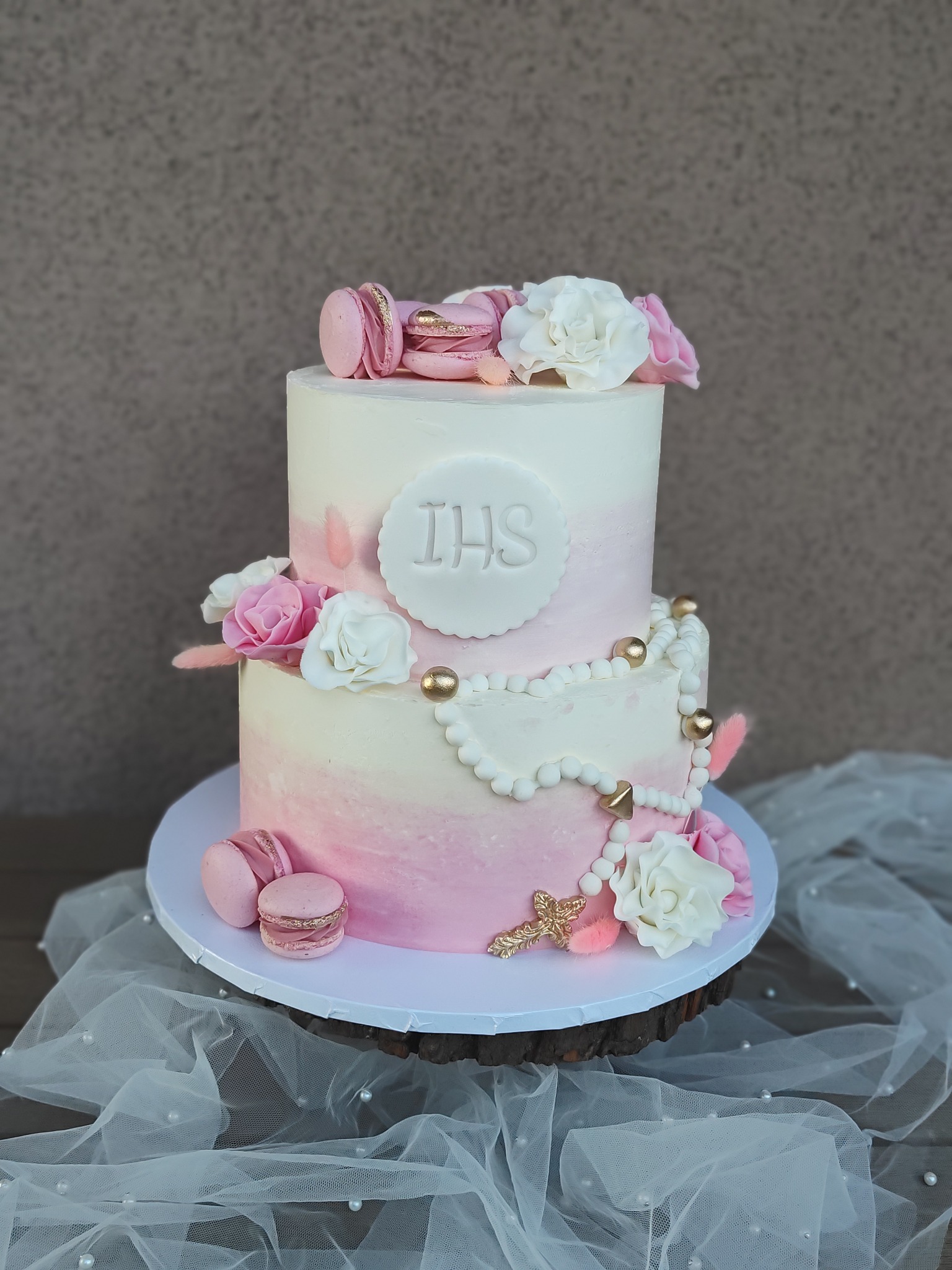 Dwupiętrowy tort ombre w odcieniach różu i bieli, dekorowany makaronikami, kwiatami i perłami, na drewnianym podeście z tiulem. Na torcie napis IHS.