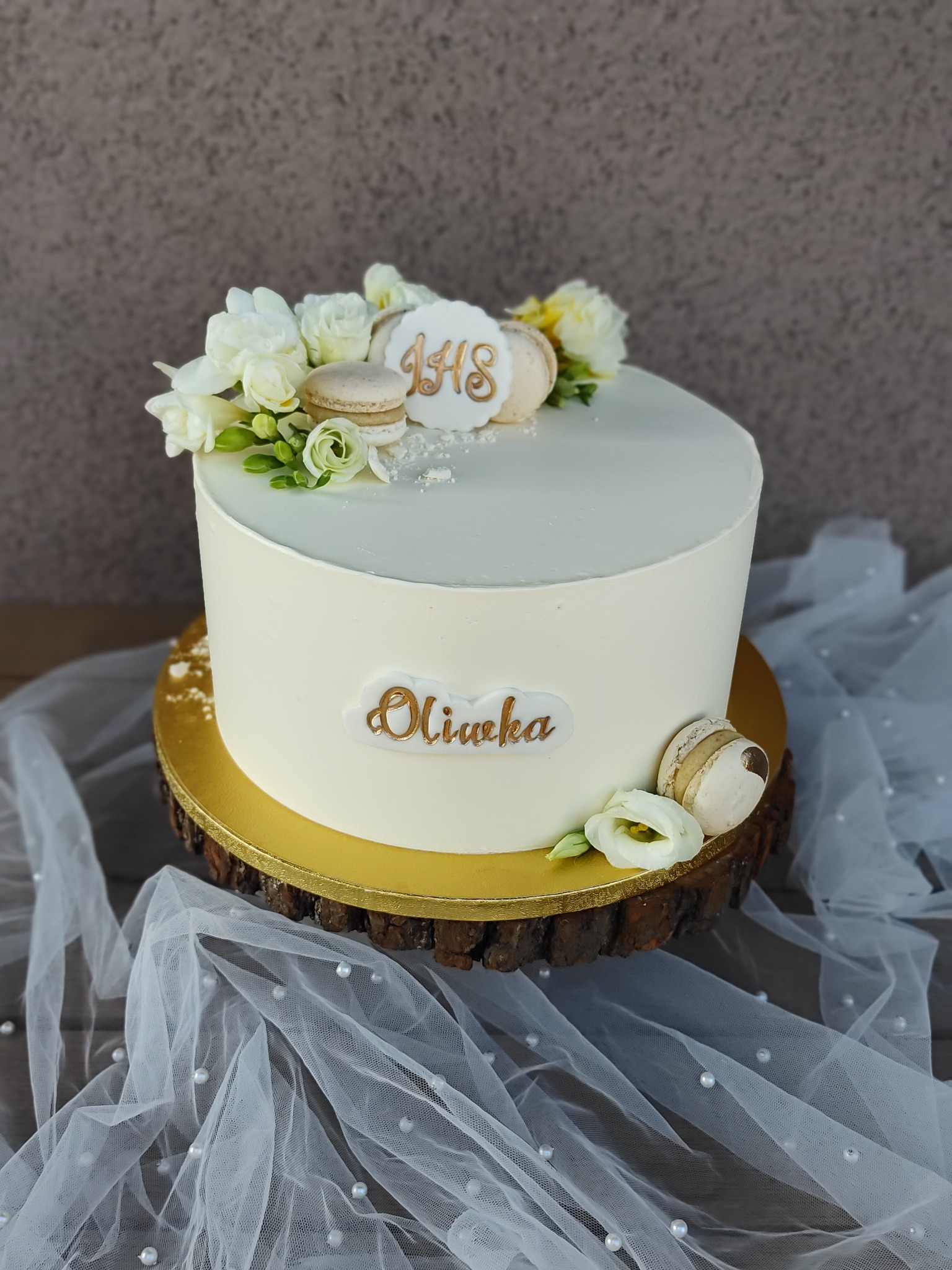 Tort urodzinowy z białym kremem, dekorowany kwiatami, makaronikami i napisem 'Oliwka' na drewnianym podeście, otoczony tiulem z perłami.
